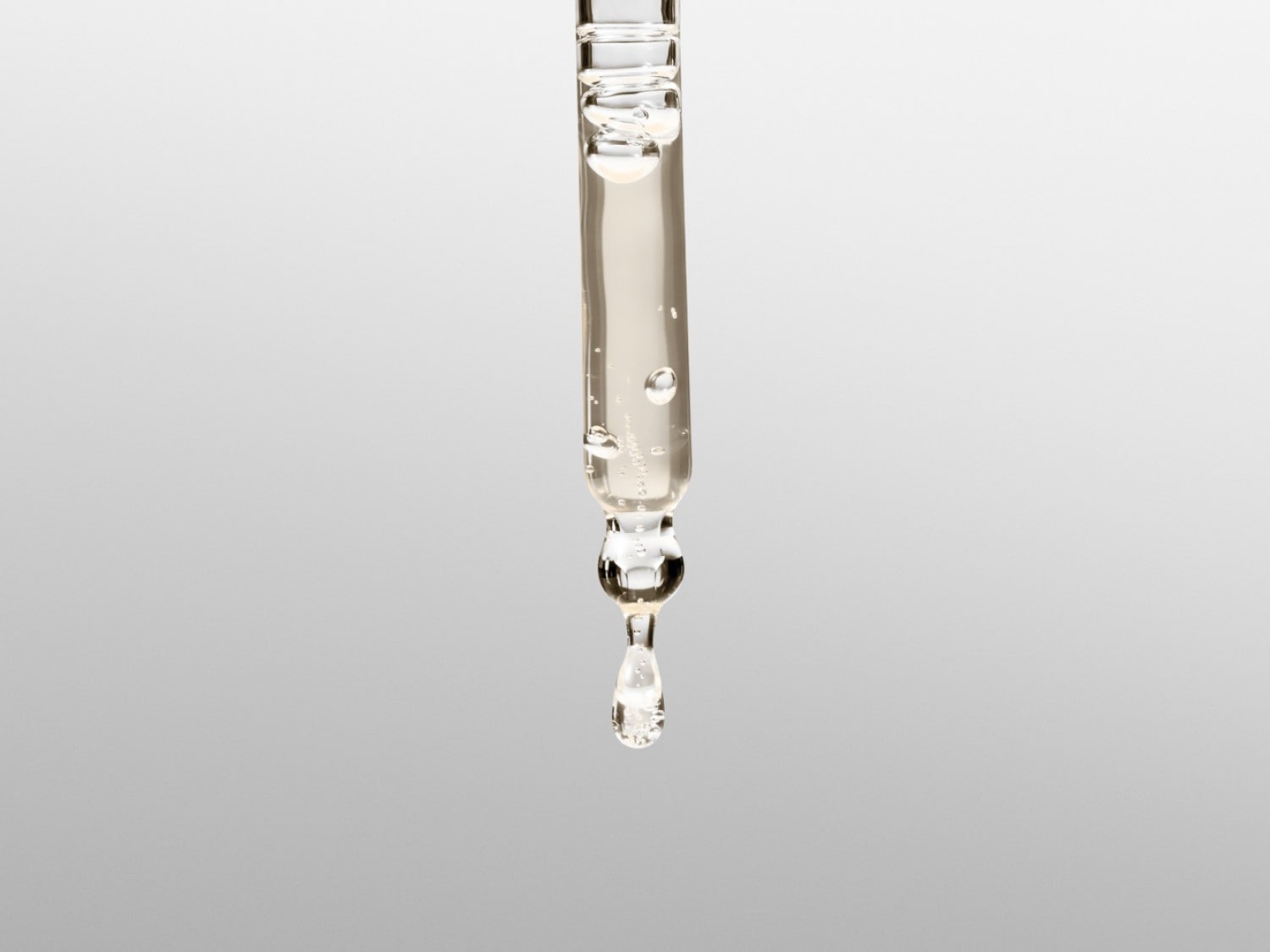 Close-up van een glazen pipet gevuld met een heldere, lichtgele vloeistof, met een enkele druppel die op het punt staat te vallen, tegen een zachte grijze achtergrond.