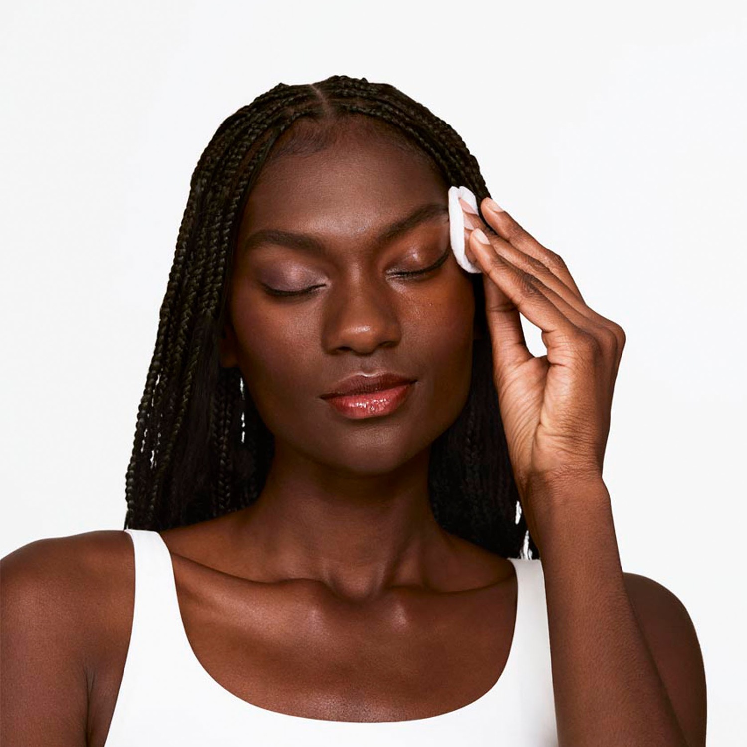 Eine Frau mit geflochtenem Haar und weißem Top reinigt ihr Gesicht sanft mit einem Wattepad.