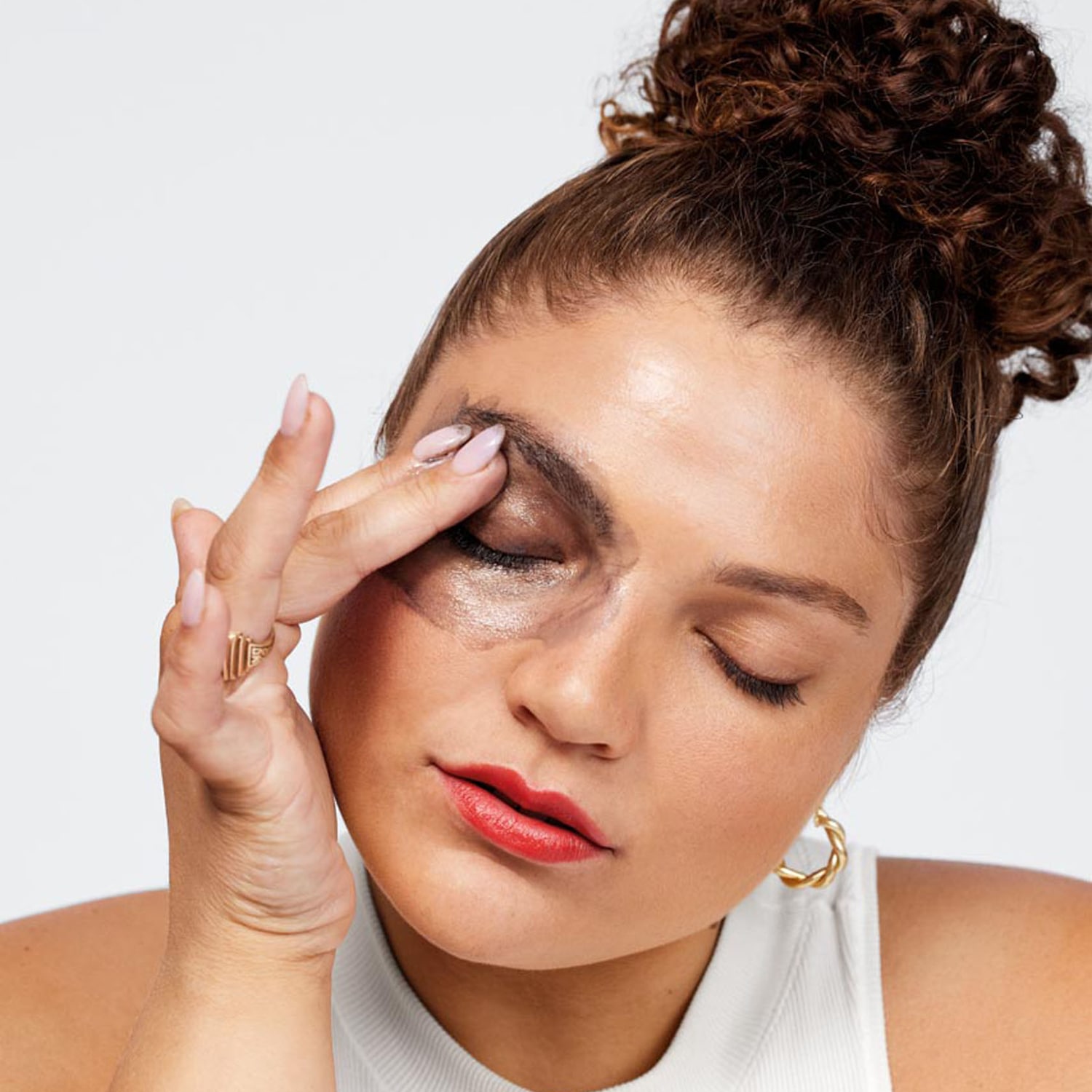 Frau mit geschlossenen Augen, die mit dem Finger grauen Lidschatten auf ihr Augenlid aufträgt, dazu roter Lippenstift und goldene Ohrringe.