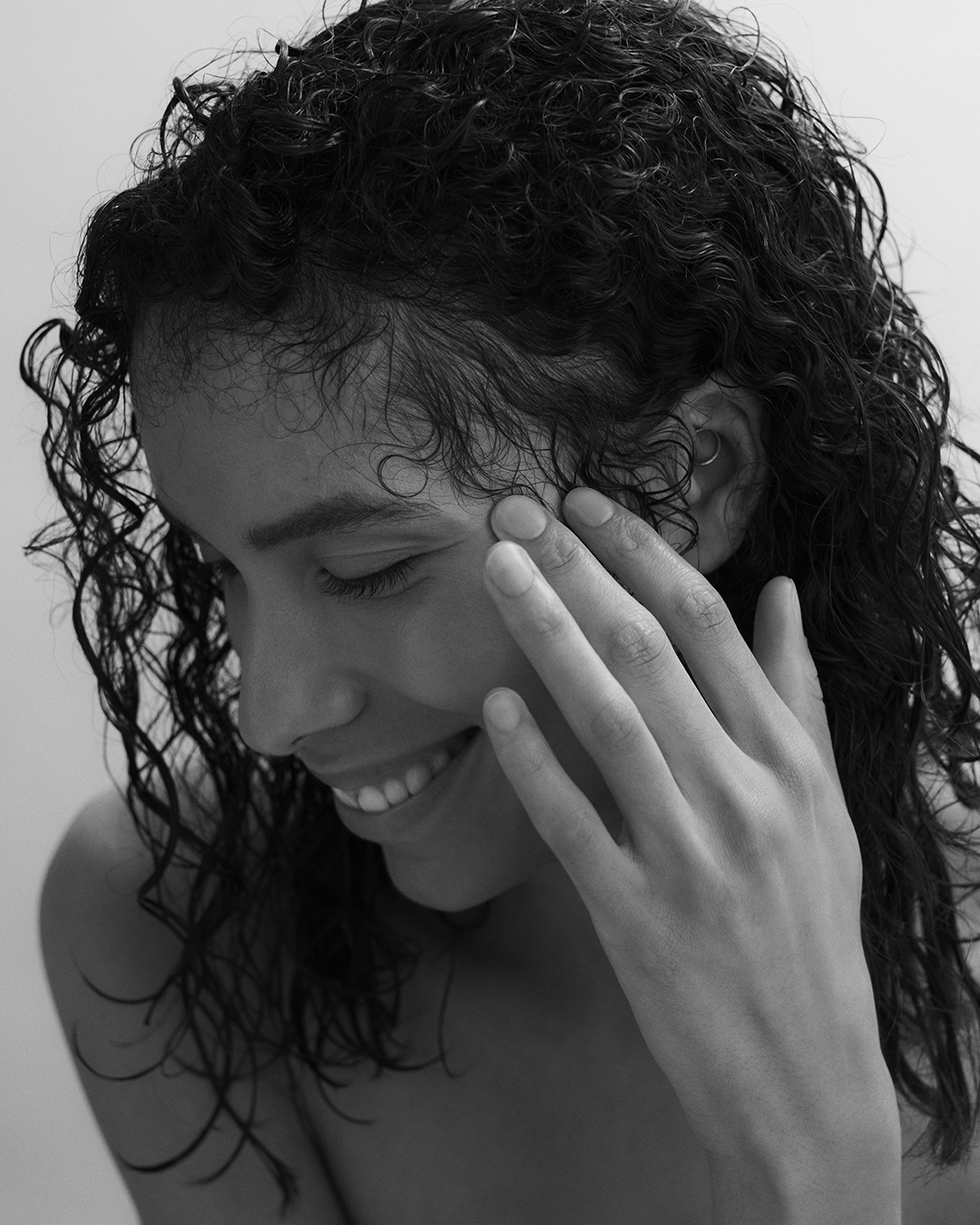 Portrait en noir et blanc d'une jeune femme souriante aux cheveux bouclés, caressant sa joue avec une main douce.