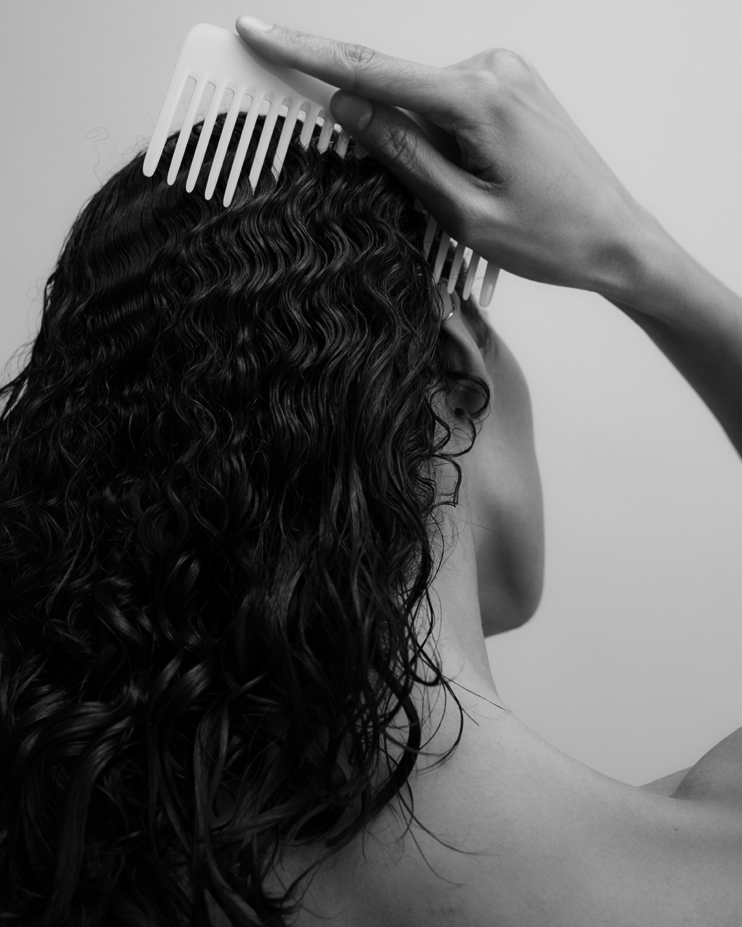 Gros plan en noir et blanc d'une personne qui se coiffe les cheveux bouclés et mouillés avec un peigne blanc à dents larges.