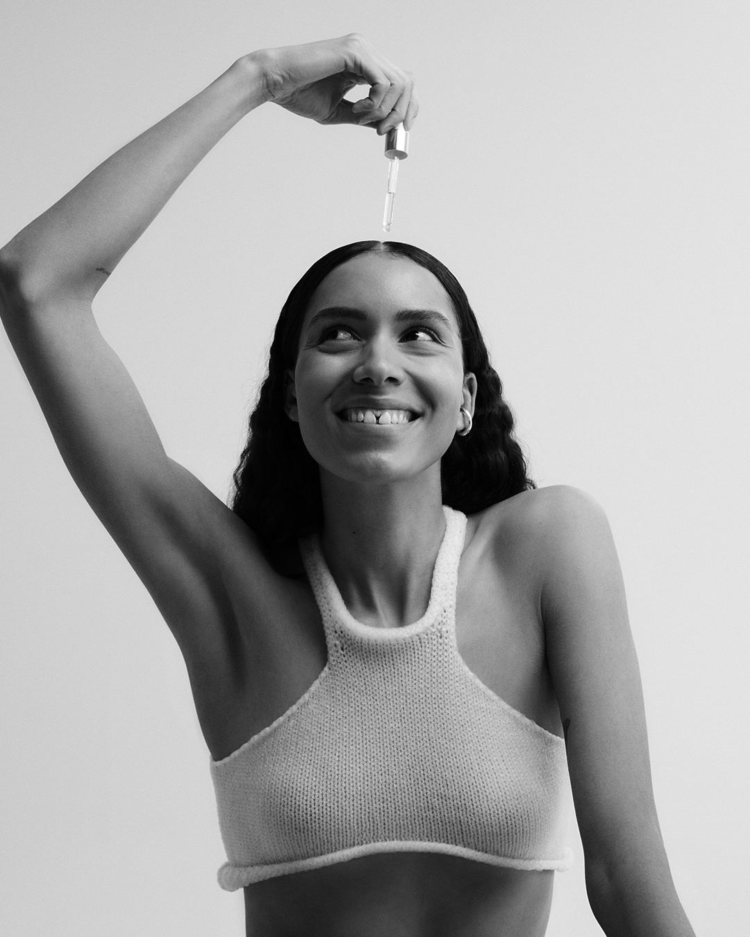 En noir et blanc, une femme souriante applique un sérum avec une pipette sur ses cheveux, vêtue d'un haut en maille.
