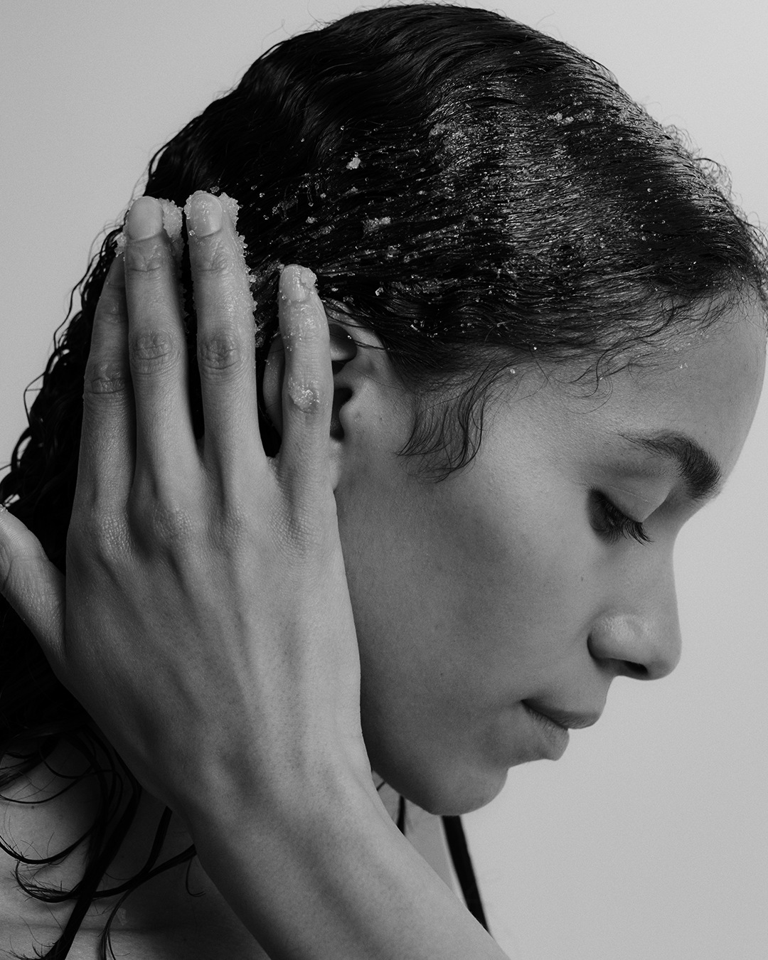 En noir et blanc, une femme applique un exfoliant granuleux sur ses cheveux mouillés, les doigts délicatement posés près de son oreille.