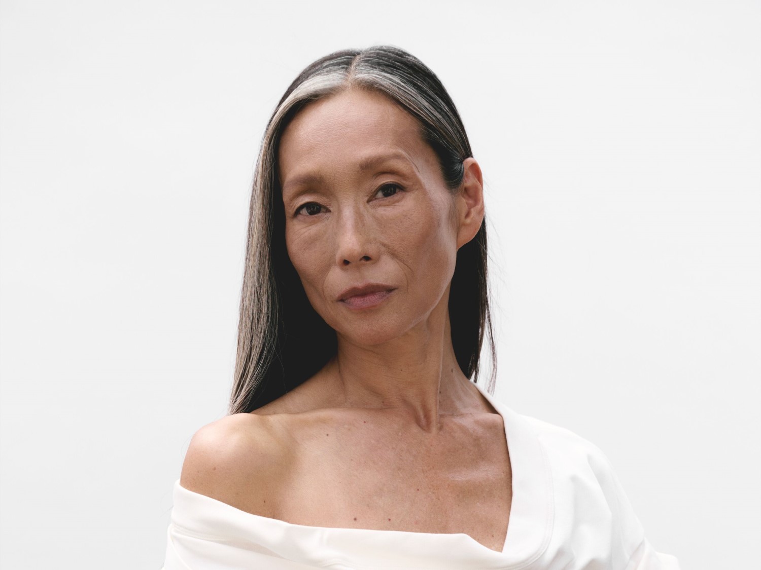 Portret van een vrouw met lang, grijs haar en een witte top, tegen een heldere achtergrond.