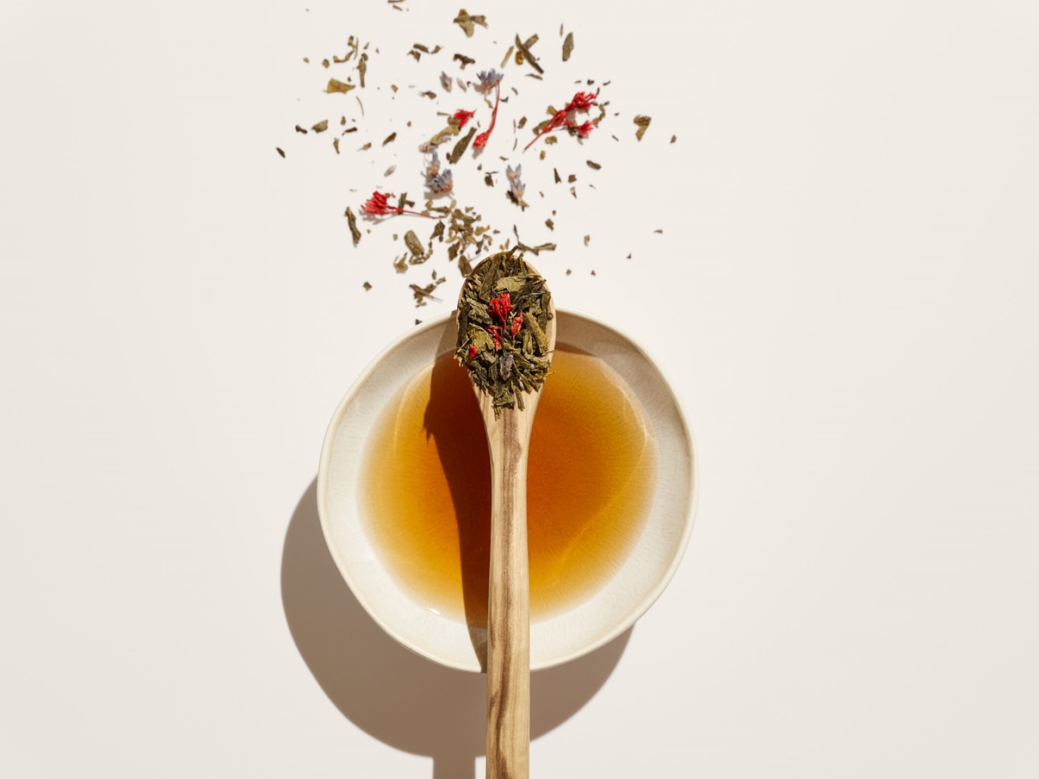 Een houten lepel gevuld met groene thee en rode bloemblaadjes rust op een schotel met thee, terwijl losse theeblaadjes en bloemblaadjes erboven zweven.