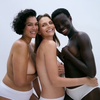 Drie vrouwen van verschillende afkomst lachen en omarmen elkaar, gekleed in witte slips, wat eenheid en diversiteit uitstraalt.