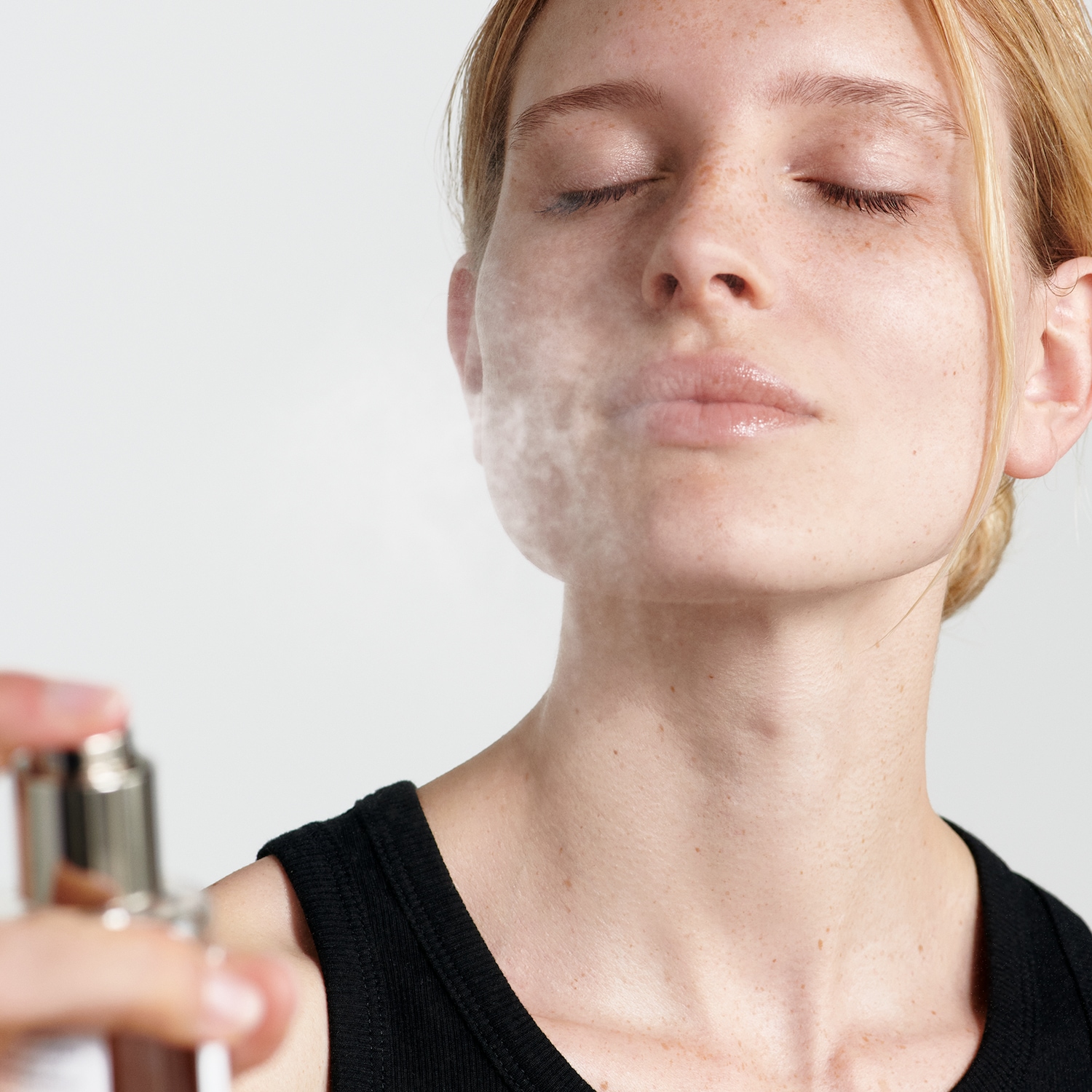 Eine Frau mit Sommersprossen schliesst die Augen, während ein feiner Nebel aus einer silbernen Sprühflasche auf ihr Gesicht gesprüht wird.