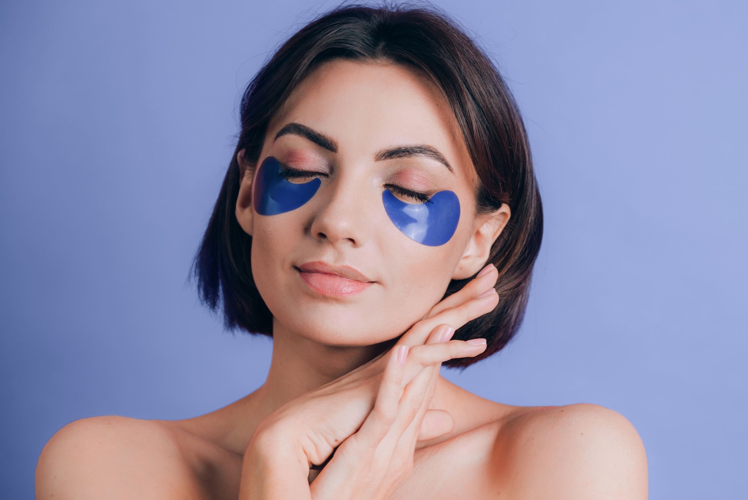 Une femme se détend avec des patchs bleus sous les yeux, les mains croisées sur la poitrine, sur un fond violet uni.