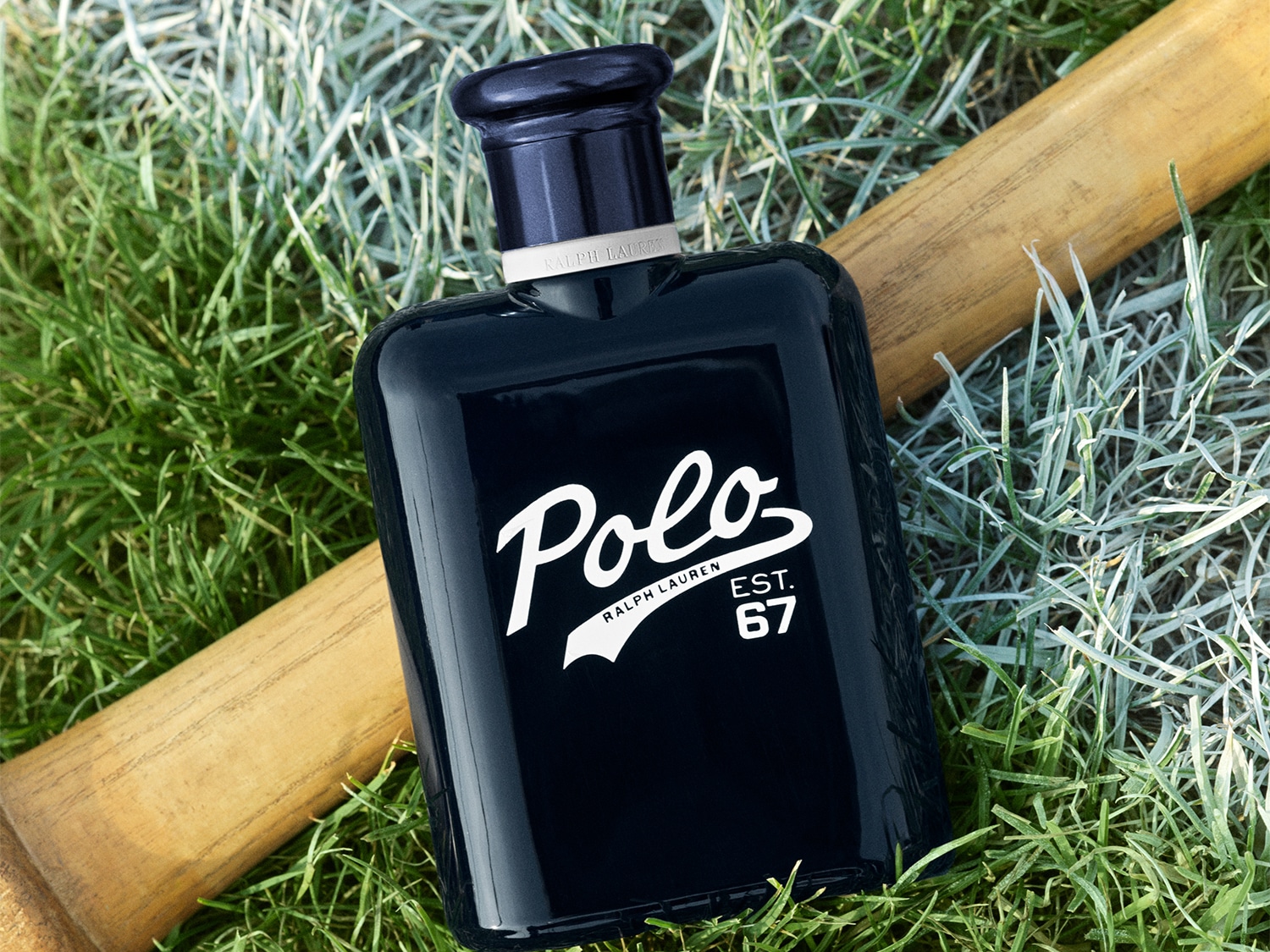 El frasco de colonia Polo Ralph Lauren EST. 67 en azul marino brillante, con letras blancas en cursiva, descansa sobre hierba verde y un bate de béisbol de madera.