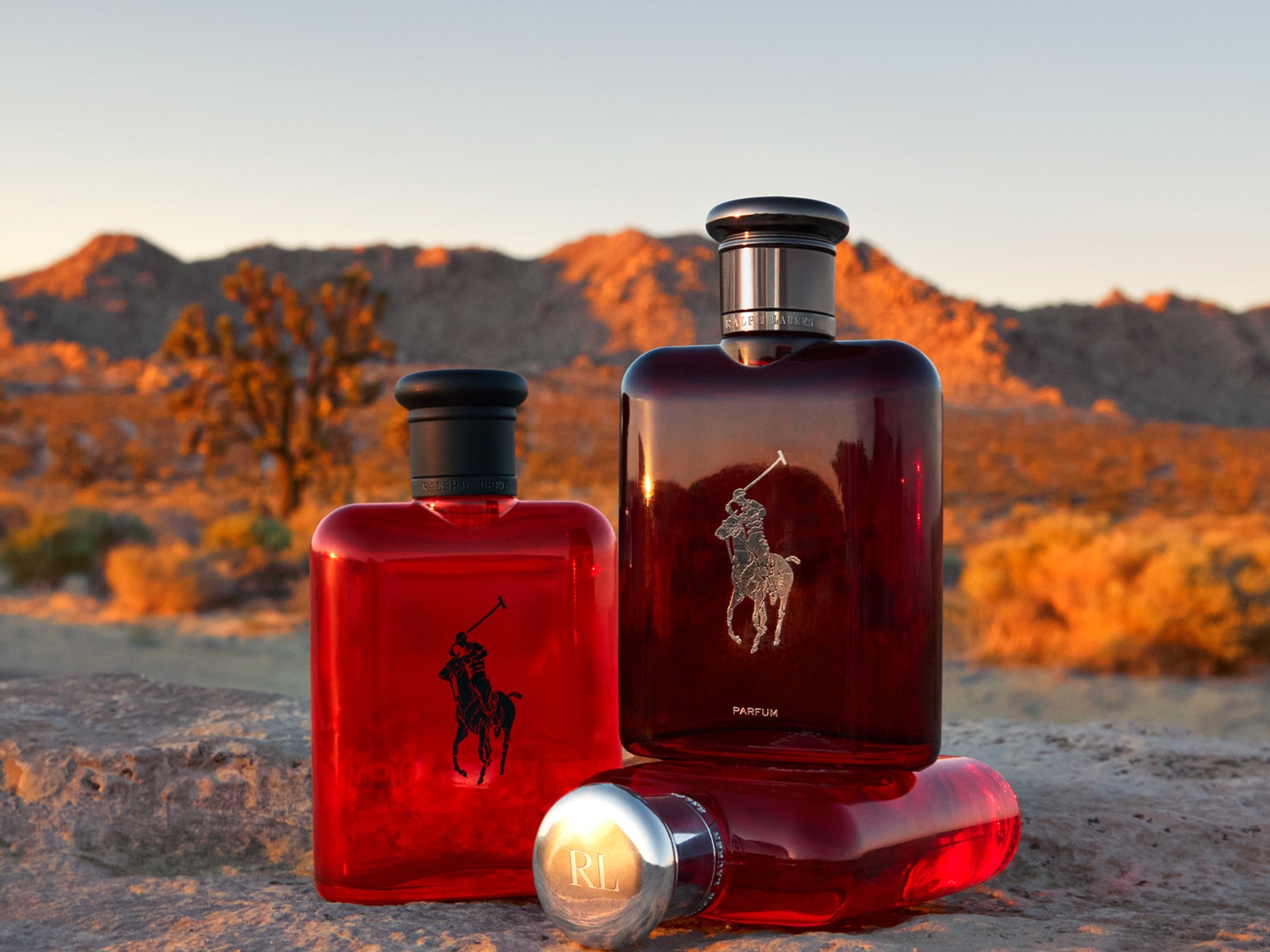 Tres frascos de perfume Ralph Lauren Polo Red se exhiben sobre una superficie rocosa, con un paisaje desértico al atardecer en el fondo, mostrando el diseño elegante y la esencia masculina de la fragancia.