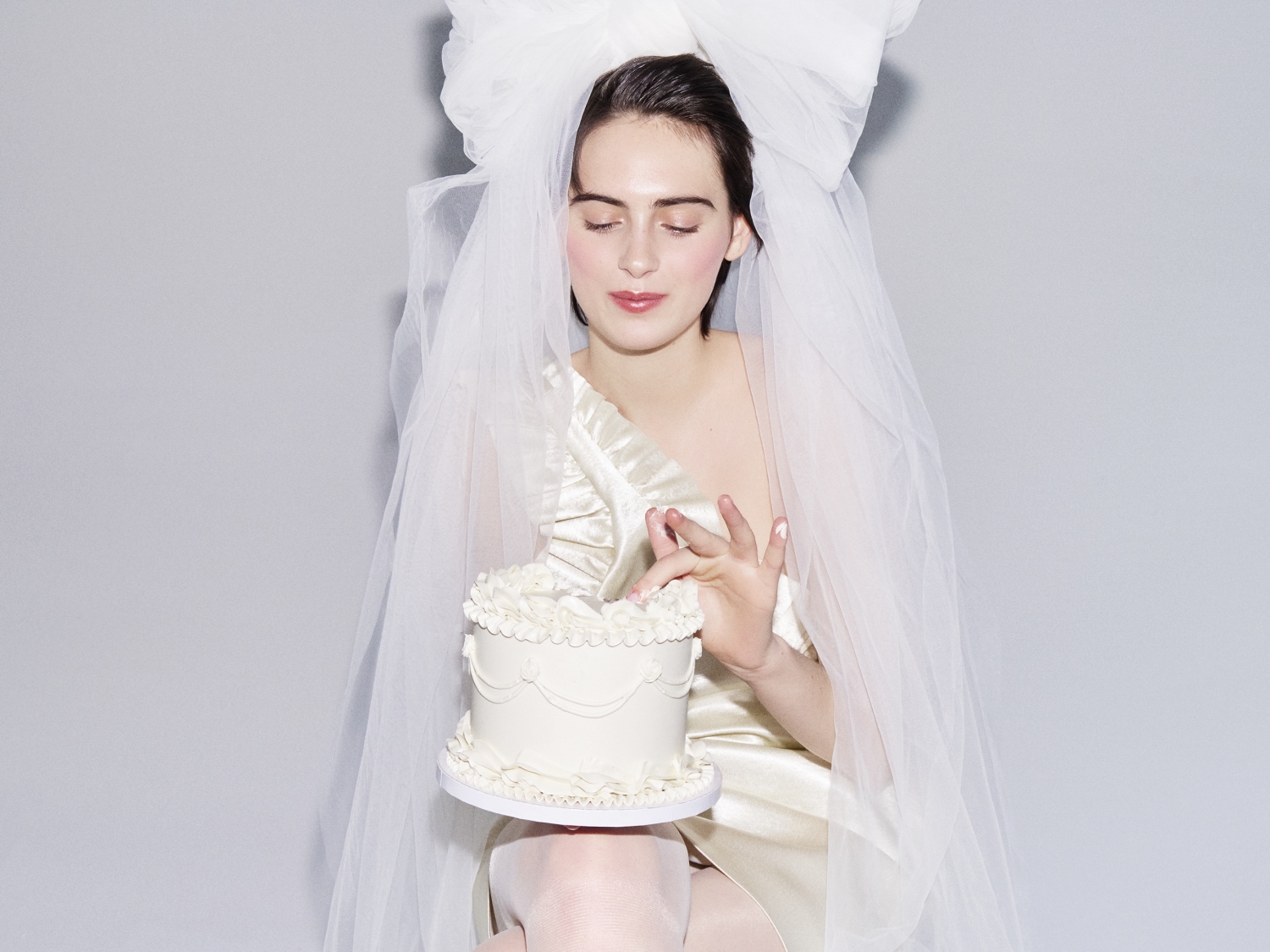 Panna młoda w welonie i sukni w kolorze kości słoniowej trzyma mały biały tort, delikatnie dotykając go palcem.