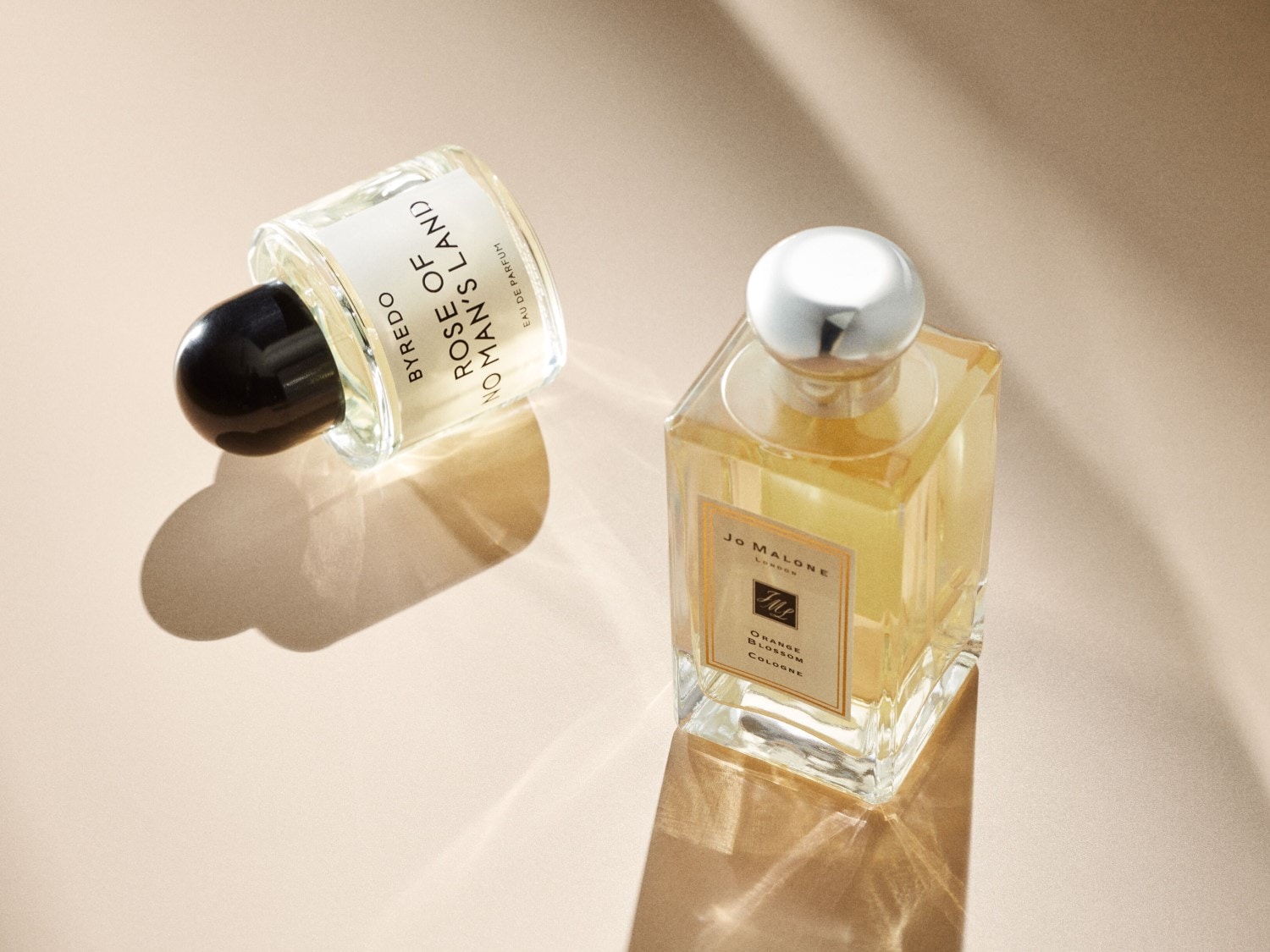 Dwa flakony perfum: Byredo Rose of No Man's Land z czarną zakrętką i Jo Malone Orange Blossom Cologne ze srebrną zakrętką, oba na beżowym tle.