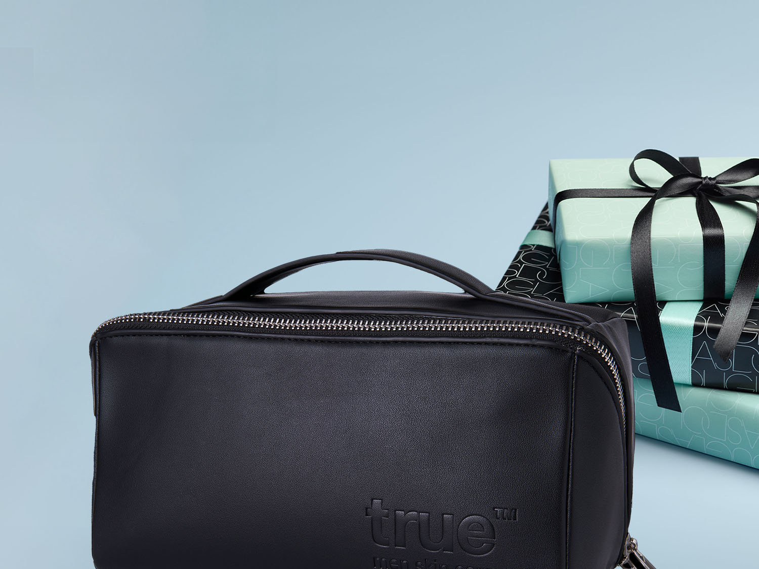 Czarna kosmetyczka z uchwytem i srebrnym zamkiem błyskawicznym stoi na tle prezentów owiniętych w czarno-turkusowy papier i przewiązanych czarną wstążką.