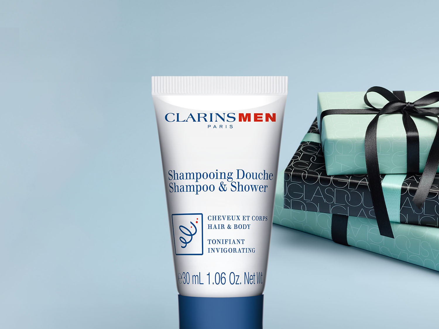 Biała tuba Clarins Men Shampooing Douche, czyli szamponu i żelu pod prysznic w jednym, z niebieskimi akcentami i tekstem, obok której leżą ozdobne prezenty.
