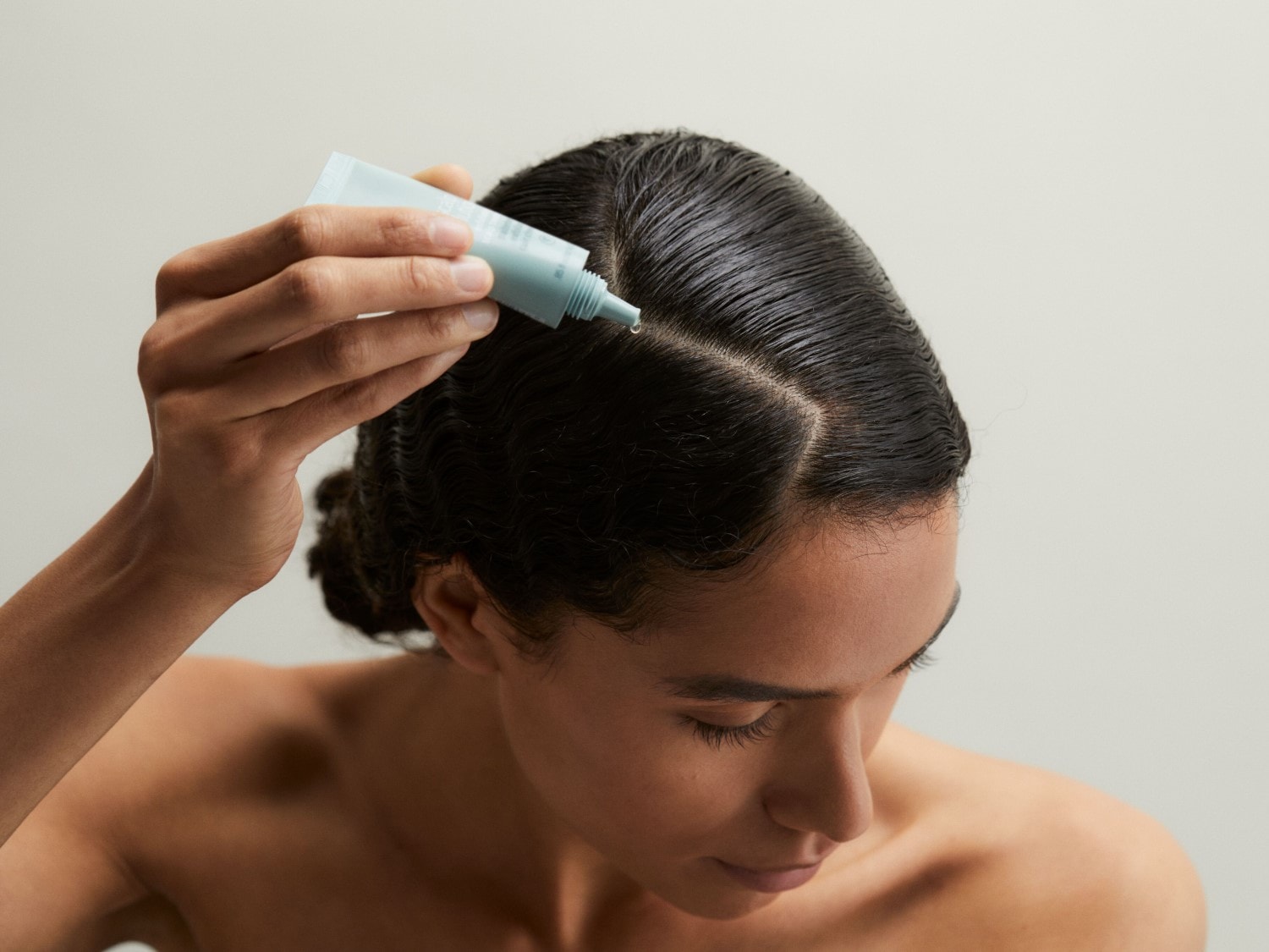 Eine Frau mit dunklen, nassen Haaren trägt eine hellblaue Tube und träufelt eine kleine Menge des Produkts auf ihre Kopfhaut.