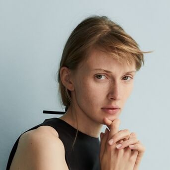 Portret van een vrouw met blond haar en een zwarte top, haar hand rustend op haar kin, tegen een lichtblauwe achtergrond.