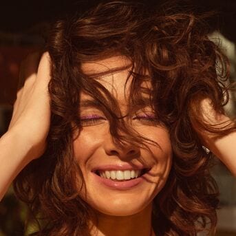 Een stralende vrouw met bruin krullend haar en paarse oogschaduw lacht met haar handen in haar haar.