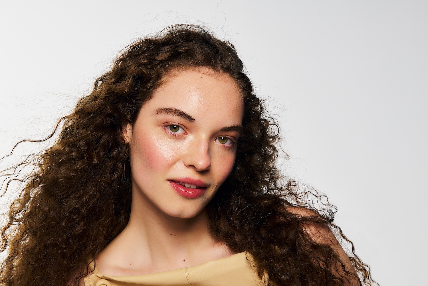 Porträt einer Frau mit lockigem, braunem Haar, zartem rosa Rouge und passendem Lippenstift, die einen hellbraunen Pullover trägt.