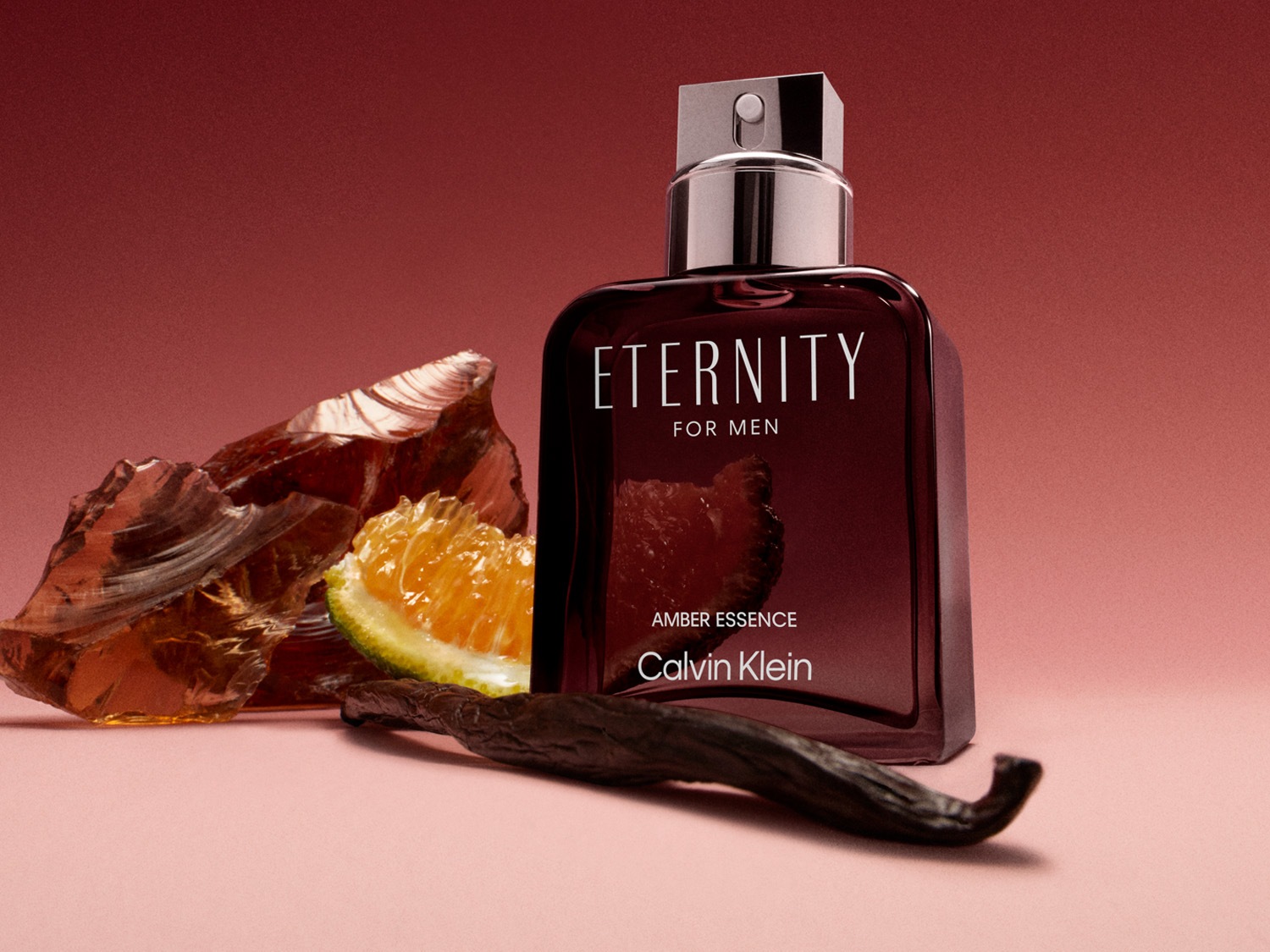Lahvička pánské vůně Calvin Klein Eternity Amber Essence s kouskem pomeranče, vanilkou a krystalem jantaru na růžovém pozadí.
