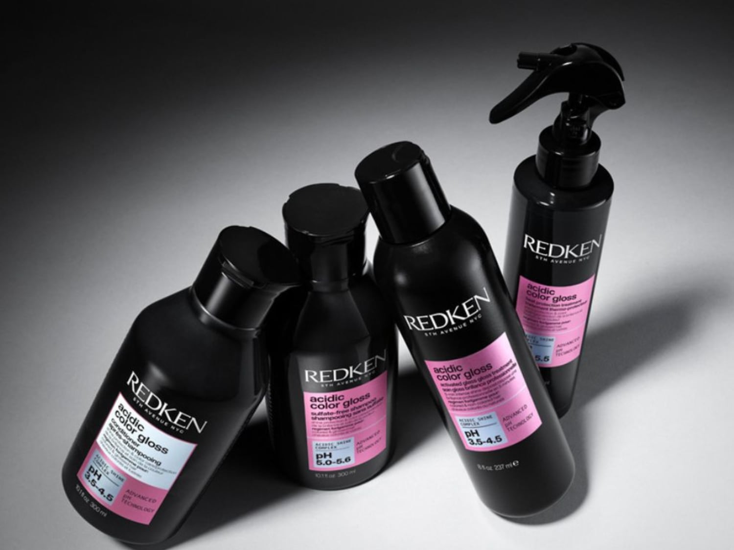 Čtyři černé lahve Redken Acidic Color Gloss s růžovými etiketami, které slibují lesk a péči o barvené vlasy.