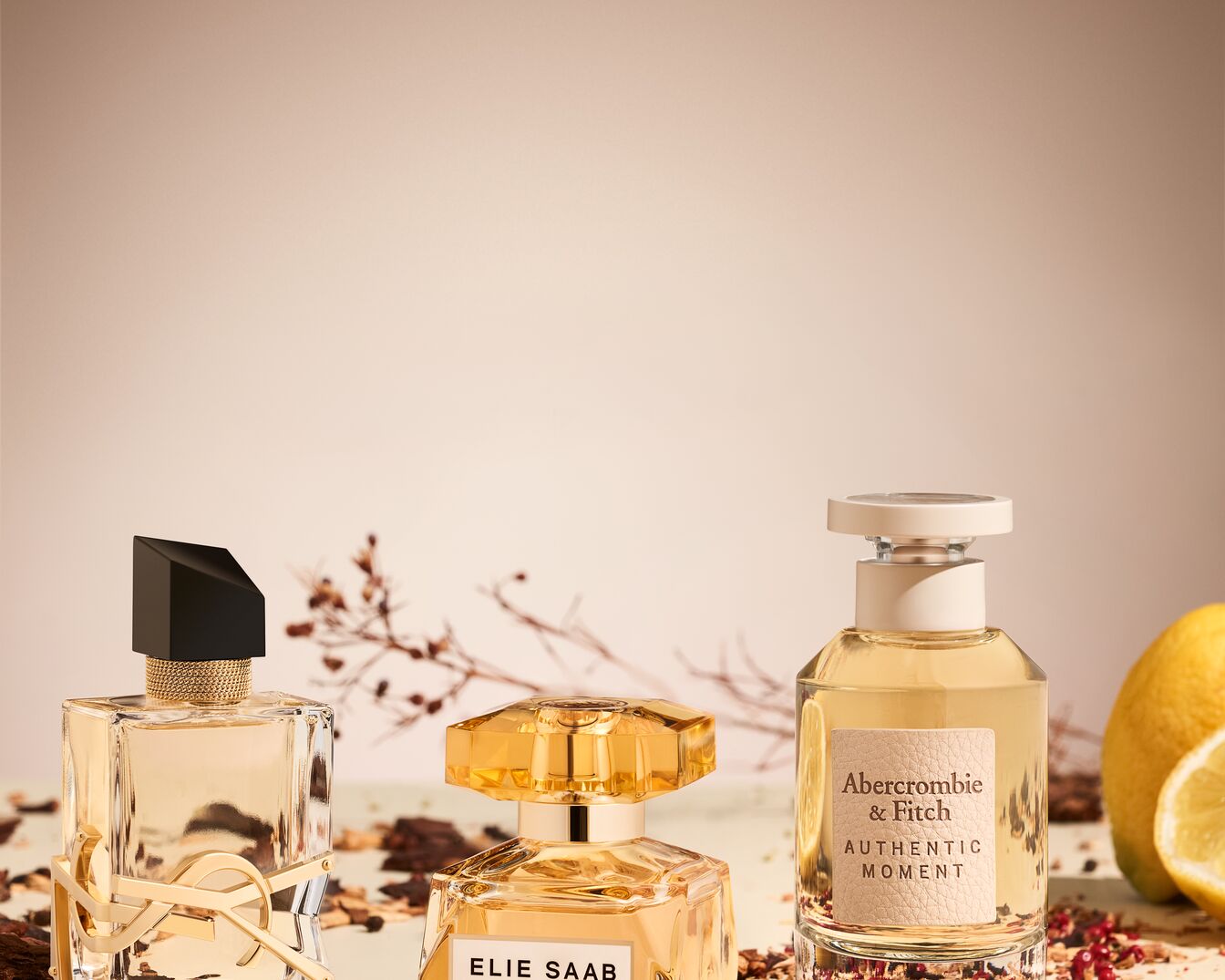 Tři flakony parfémů různých značek, včetně YSL, Elie Saab a Abercrombie & Fitch, s teplými, zlatavými tóny a přírodními dekoracemi.