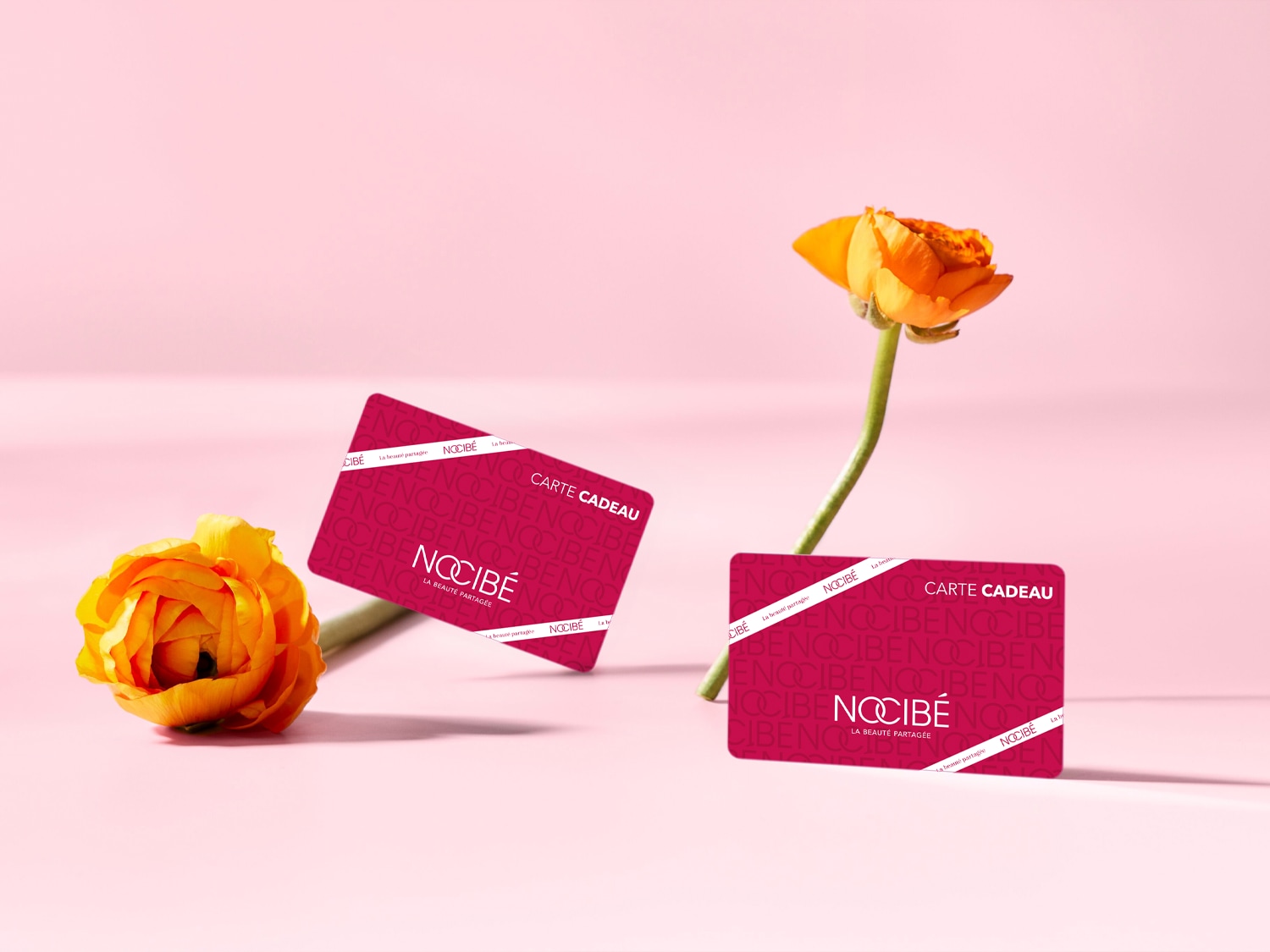 Deux cartes cadeaux Nocibé bordeaux sont présentées avec des fleurs orange sur un fond rose, offrant un cadeau de beauté partagée.