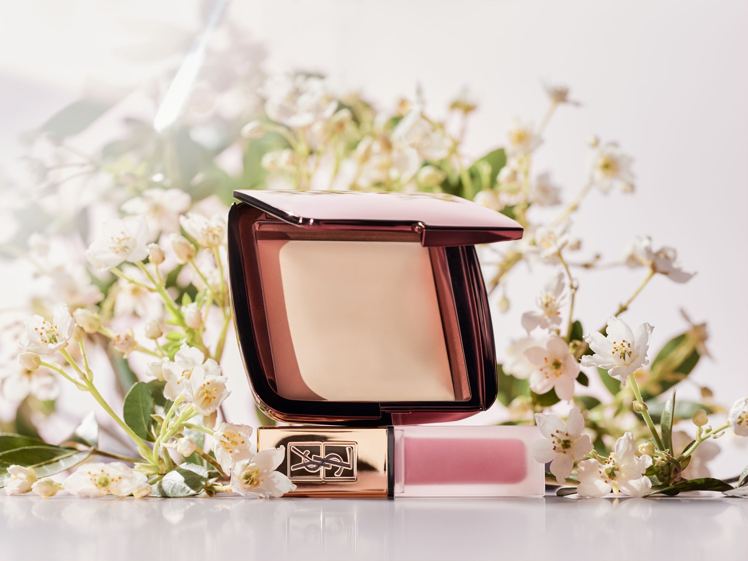 Poudre compacte dans un boîtier rose et marron, posée sur un rouge à lèvres rose pâle, entourée de délicates fleurs blanches.