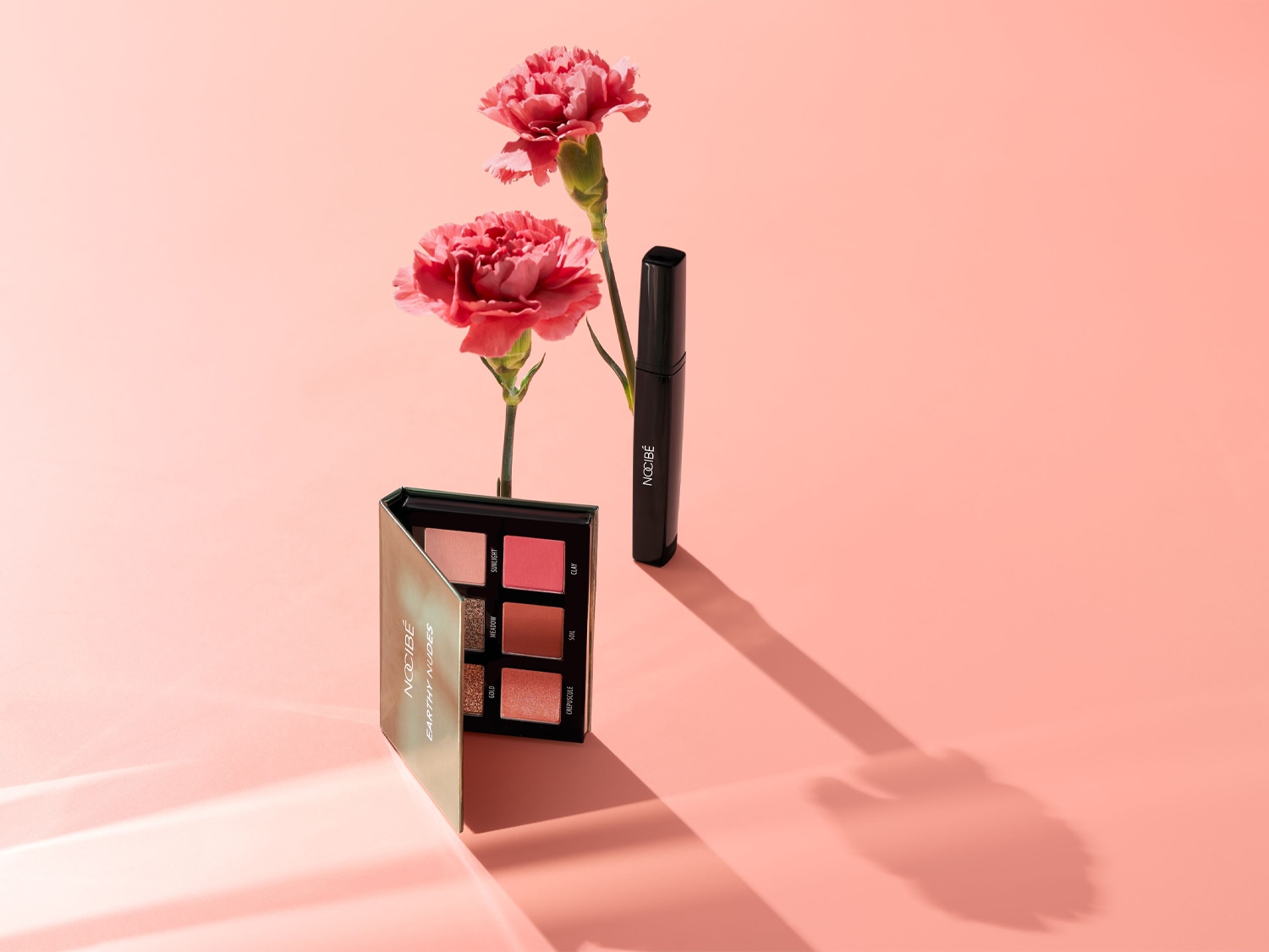 Palette de fards à paupières Nocibé Earthy Nudes aux tons chauds et mascara noir, présentés avec des œillets roses sur un fond pêche.