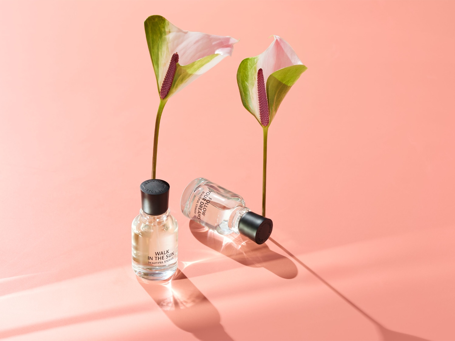 Deux parfums reposent sur un fond rose pêche, accompagnés de deux fleurs d'anthurium délicates, créant une ambiance à la fois fraîche et élégante.