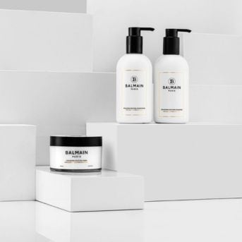 Dva bílé flakony a kelímek vlasové kosmetiky Balmain Paris s černými detaily na bílém pozadí.