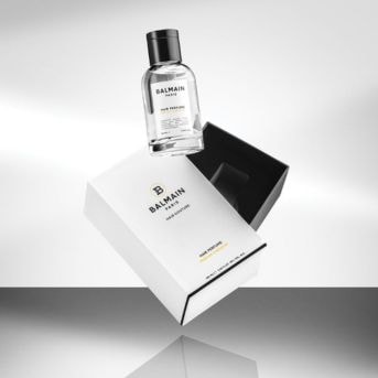 Průhledná skleněná lahvička Balmain Hair Perfume se vznáší nad bílou krabičkou s černým vnitřkem, elegantní a luxusní.