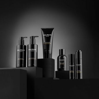 Řada černých lahviček a tub s produkty Balmain Paris pro péči o vlasy a pleť, elegantně uspořádaná na černých podstavcích.