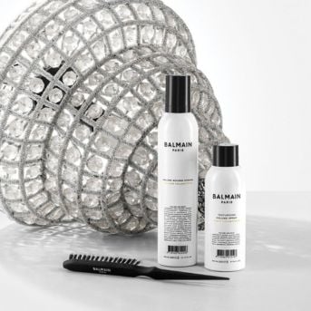 Dva bílé produkty Balmain Paris s černými víčky a kartáč na vlasy leží před stříbrnou dekorativní koulí.