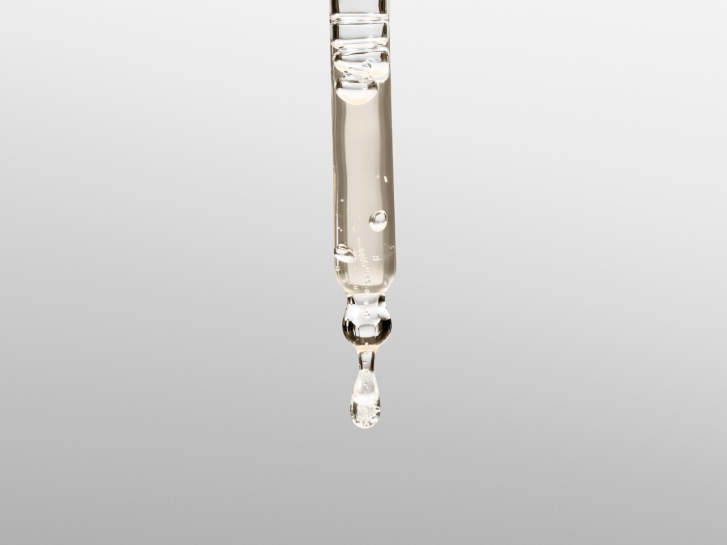 Close-up van een glazen pipet gevuld met een heldere, lichtgele vloeistof, met een enkele druppel die op het punt staat te vallen, tegen een zachte grijze achtergrond.