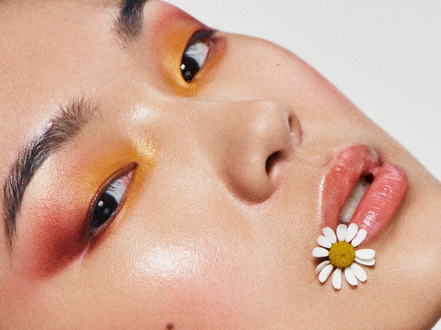 Nahaufnahme eines Gesichts mit orangefarbenem Lidschatten und einem Gänseblümchen zwischen den Lippen, das einen frischen und natürlichen Look erzeugt.