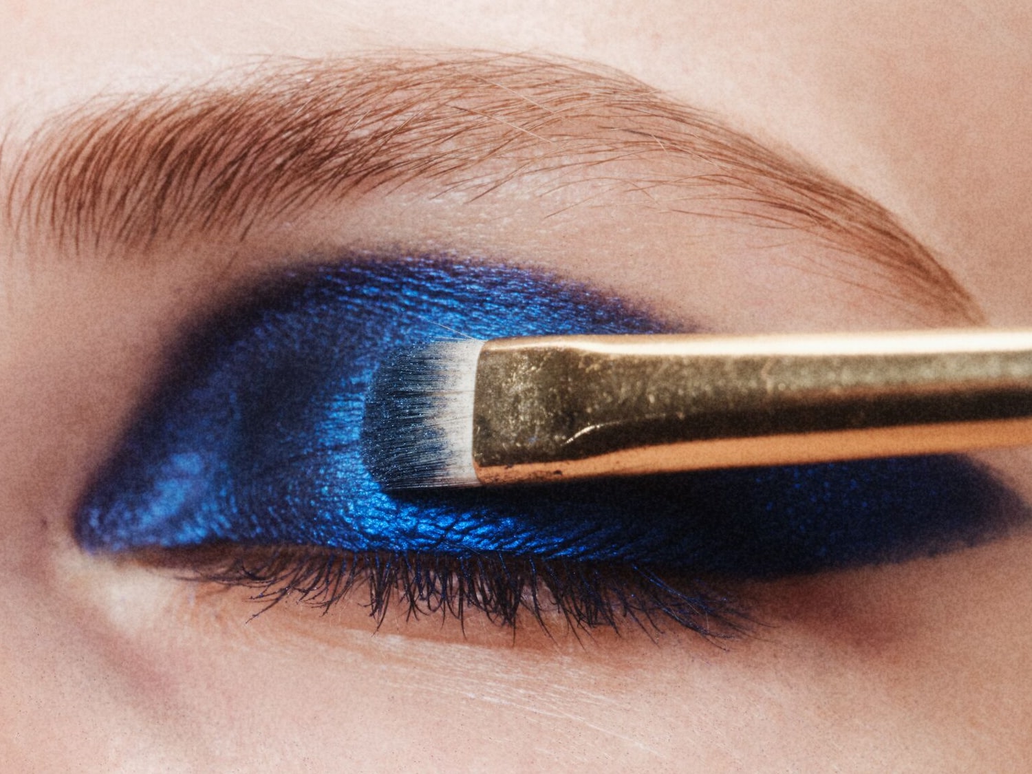 Nahaufnahme eines Auges mit blauem Lidschatten, der mit einem goldenen Pinsel aufgetragen wird, um einen dramatischen und farbenfrohen Look zu erzielen.