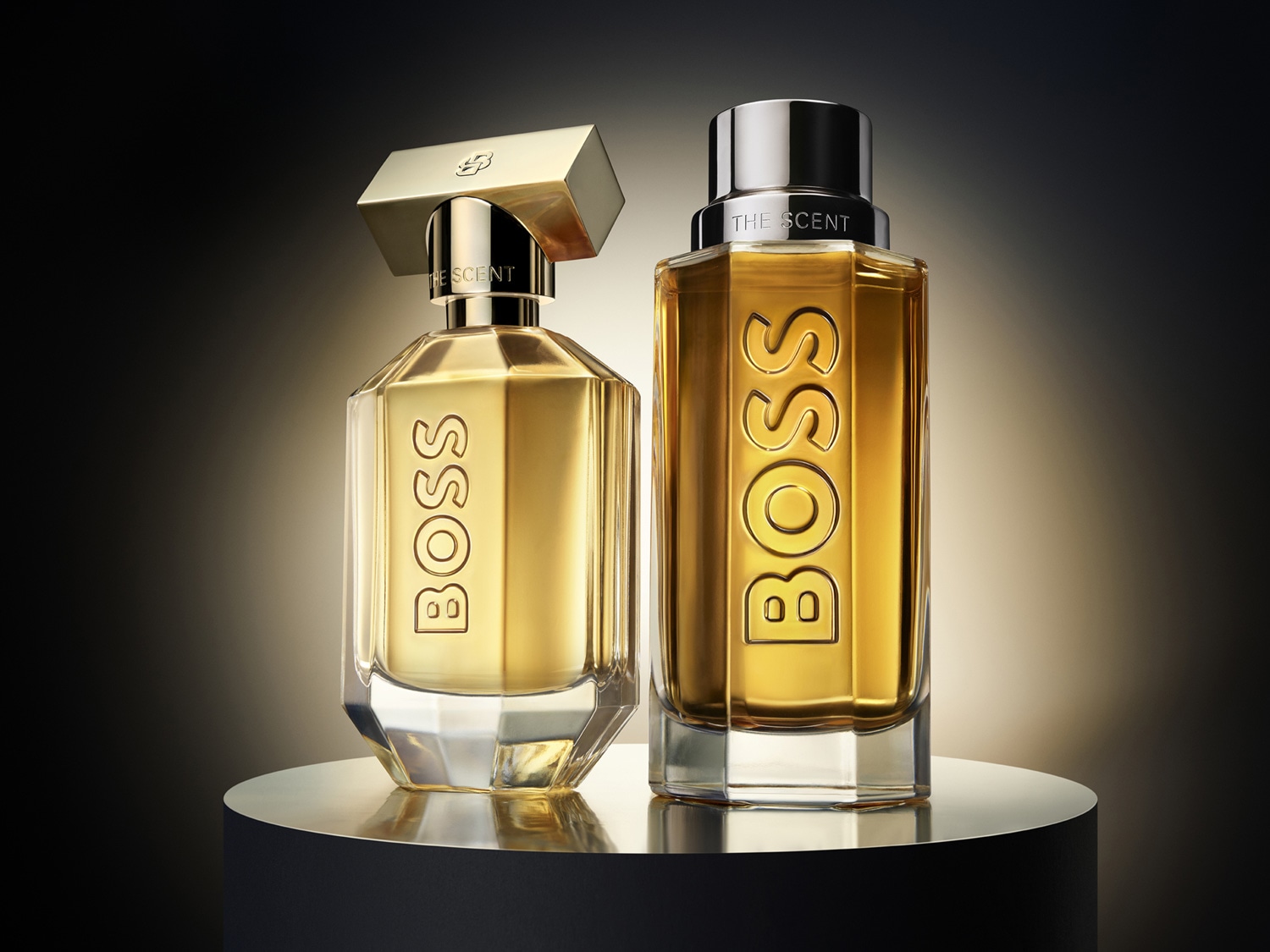 Dva flakóny vône BOSS The Scent, jeden s hranatým zlatým vrchnákom a druhý s kovovým strieborným vrchnákom, stoja vedľa seba na lesklom čiernom podstavci.