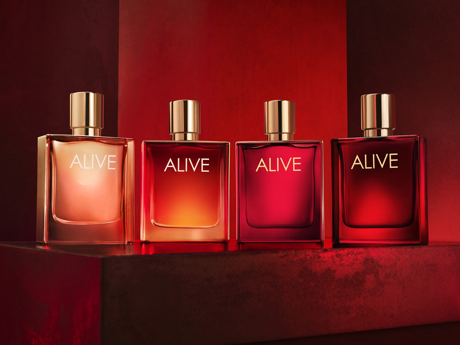 Štyri flakóny vône ALIVE v odtieňoch od svetlej po tmavočervenú, každý s nápisom ALIVE a zlatým vrchnákom, vytvárajúce teplý a luxusný dojem.