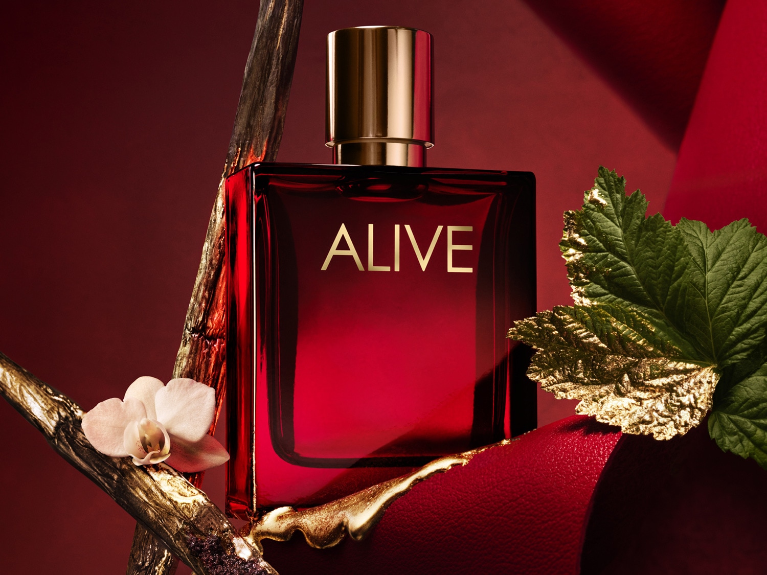 Čtvercový flakon parfému Alive v červeném provedení se zlatým uzávěrem a nápisem, doplněný o květinu a list.