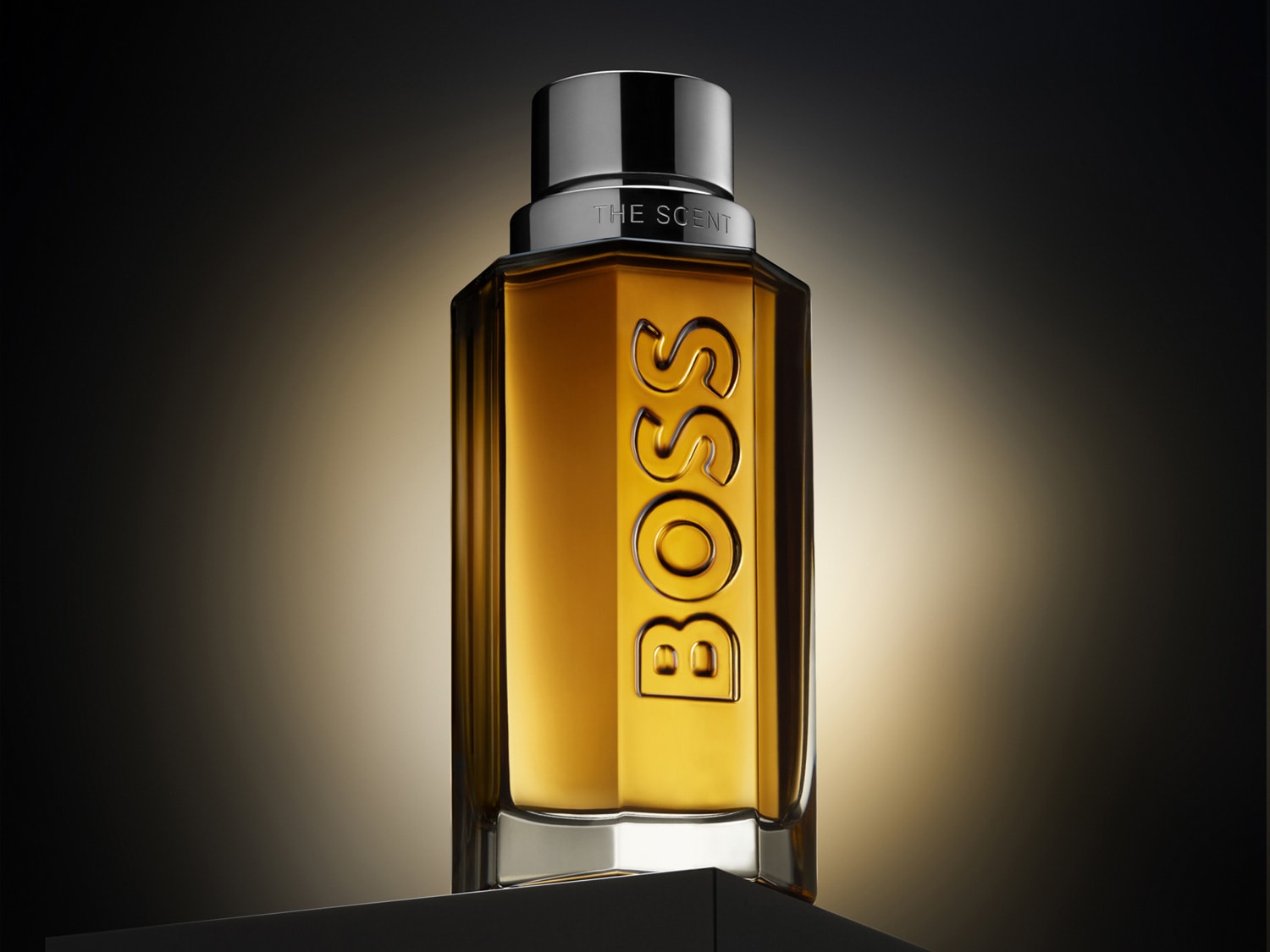 Lahvička parfému BOSS The Scent s hnědozlatou tekutinou a stříbrným uzávěrem, na které je vyryto logo BOSS.