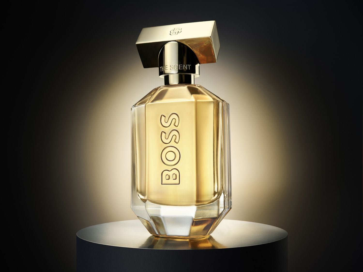 Zlatavá vůně BOSS The Scent v elegantním skleněném flakonu s výrazným zlatým uzávěrem, umístěná na lesklém podstavci.