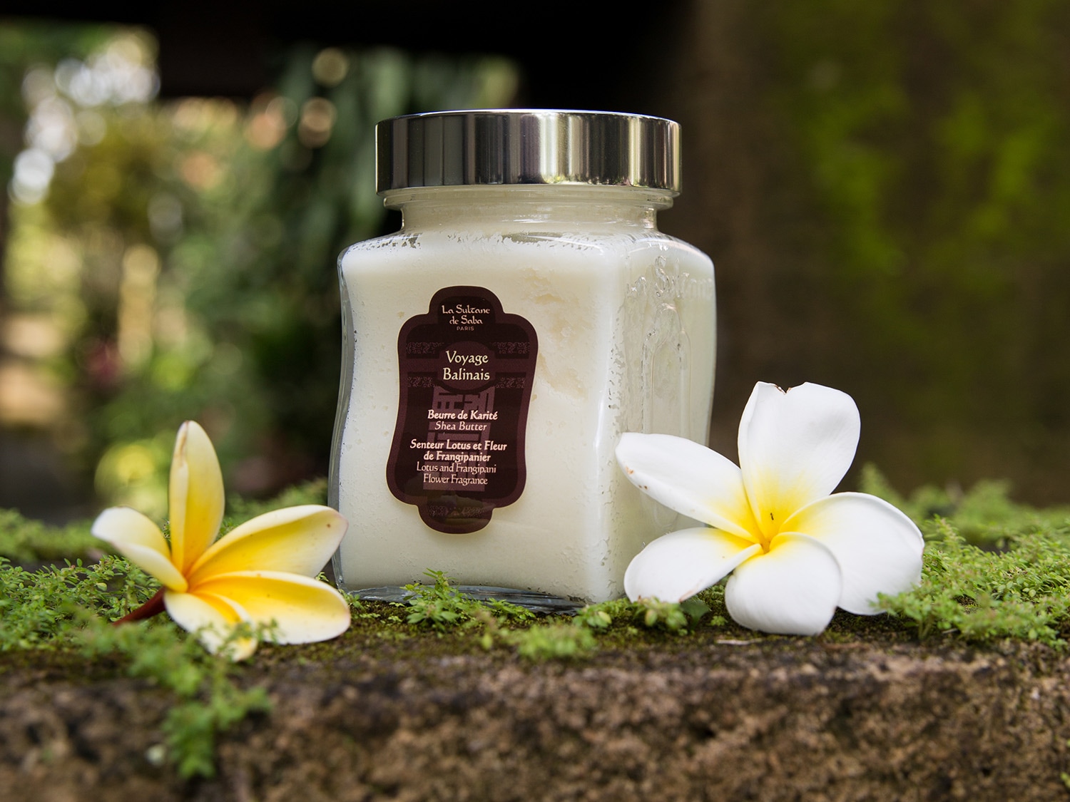 Krem do ciała La Sultane de Saba Voyage Balinais o zapachu lotosu i kwiatu frangipani, zamknięty w szklanym słoiczku z metalową nakrętką, otoczony kwiatami.