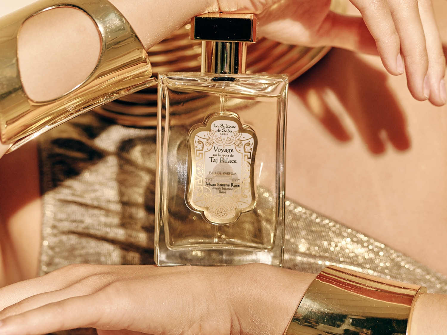 Przezroczysty flakon perfum La Sultane de Saba Voyage sur la route du Taj Palace Eau de Parfum, trzymany w dłoniach ozdobionych złotymi bransoletami, emanuje luksusem i egzotyką.