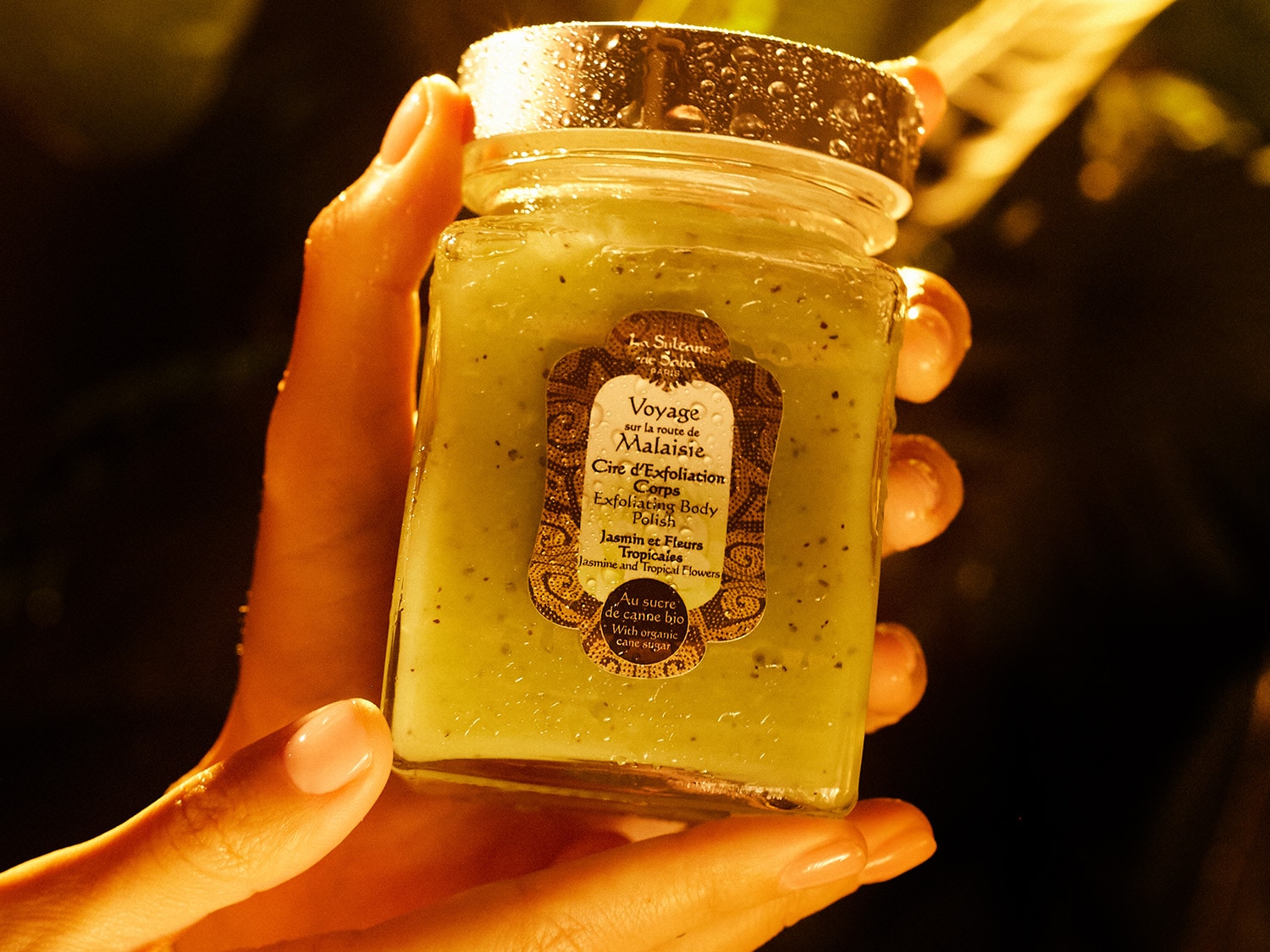Dłoń trzyma słoik z pilingiem do ciała La Sultane de Saba Voyage sur la route de Malaisie o zapachu jaśminu i kwiatów tropikalnych.