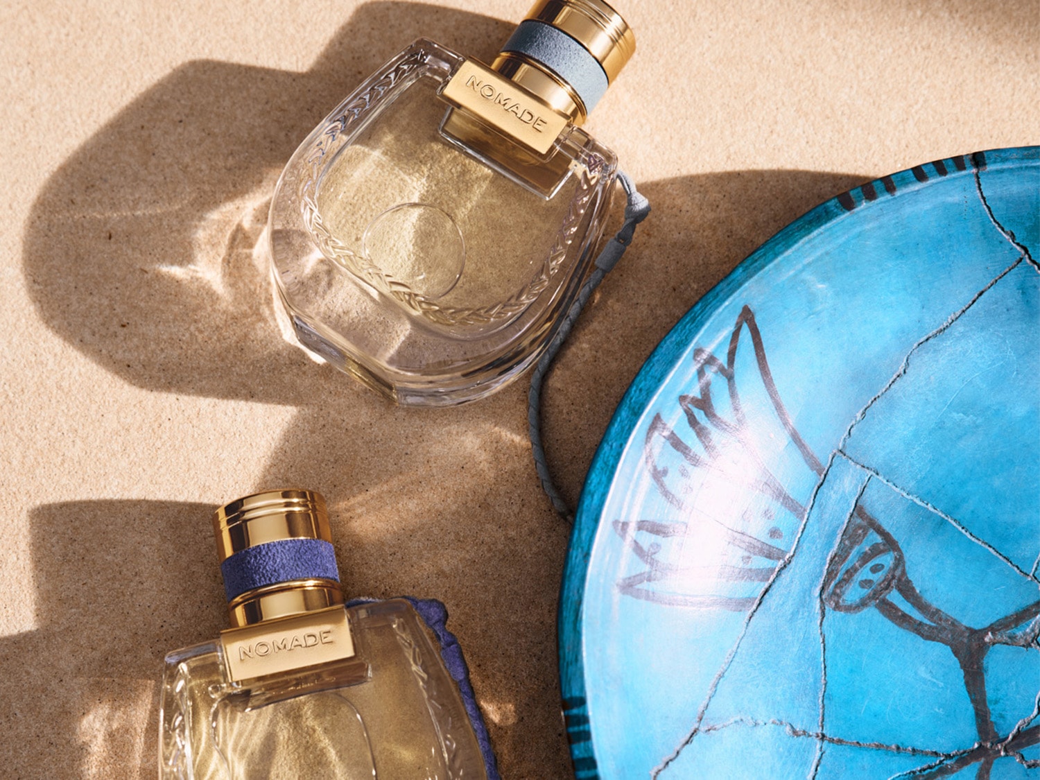 Dva flakony parfému Nomade leží na písku vedle modrého talíře s kresbou, zlatavá tekutina uvnitř skleněných lahviček slibuje vůni dobrodružství a svobody.