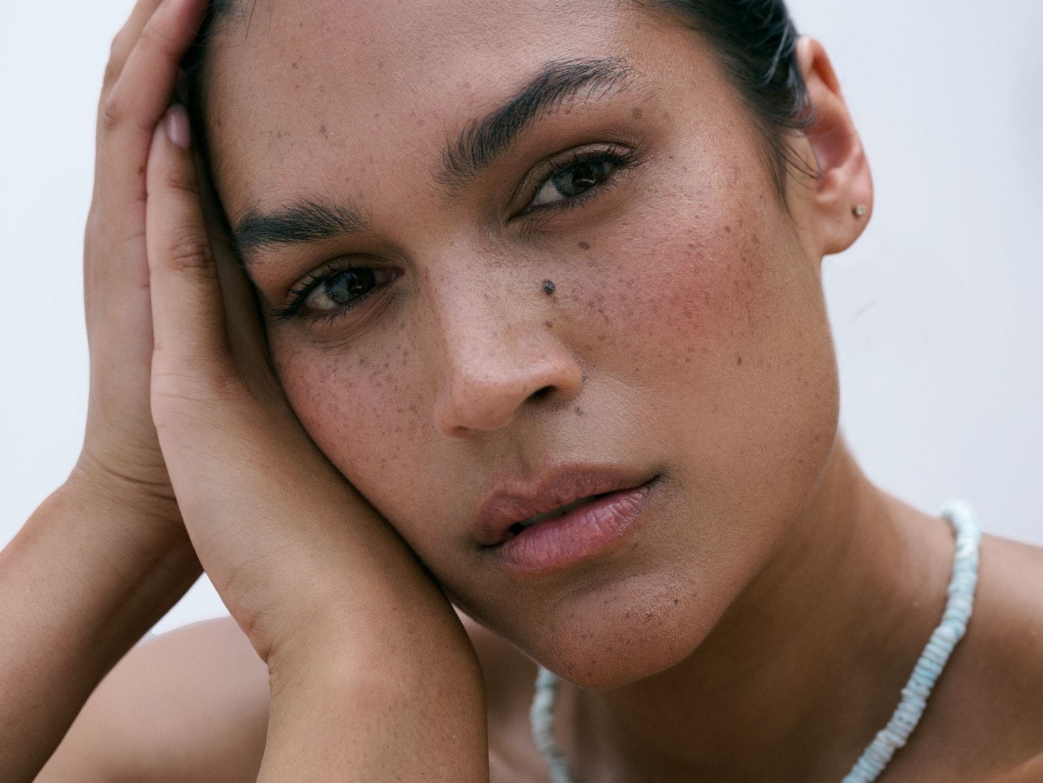 Nahaufnahme einer Frau mit Sommersprossen, die ihre Hände an ihr Gesicht legt und eine hellblaue Halskette trägt.