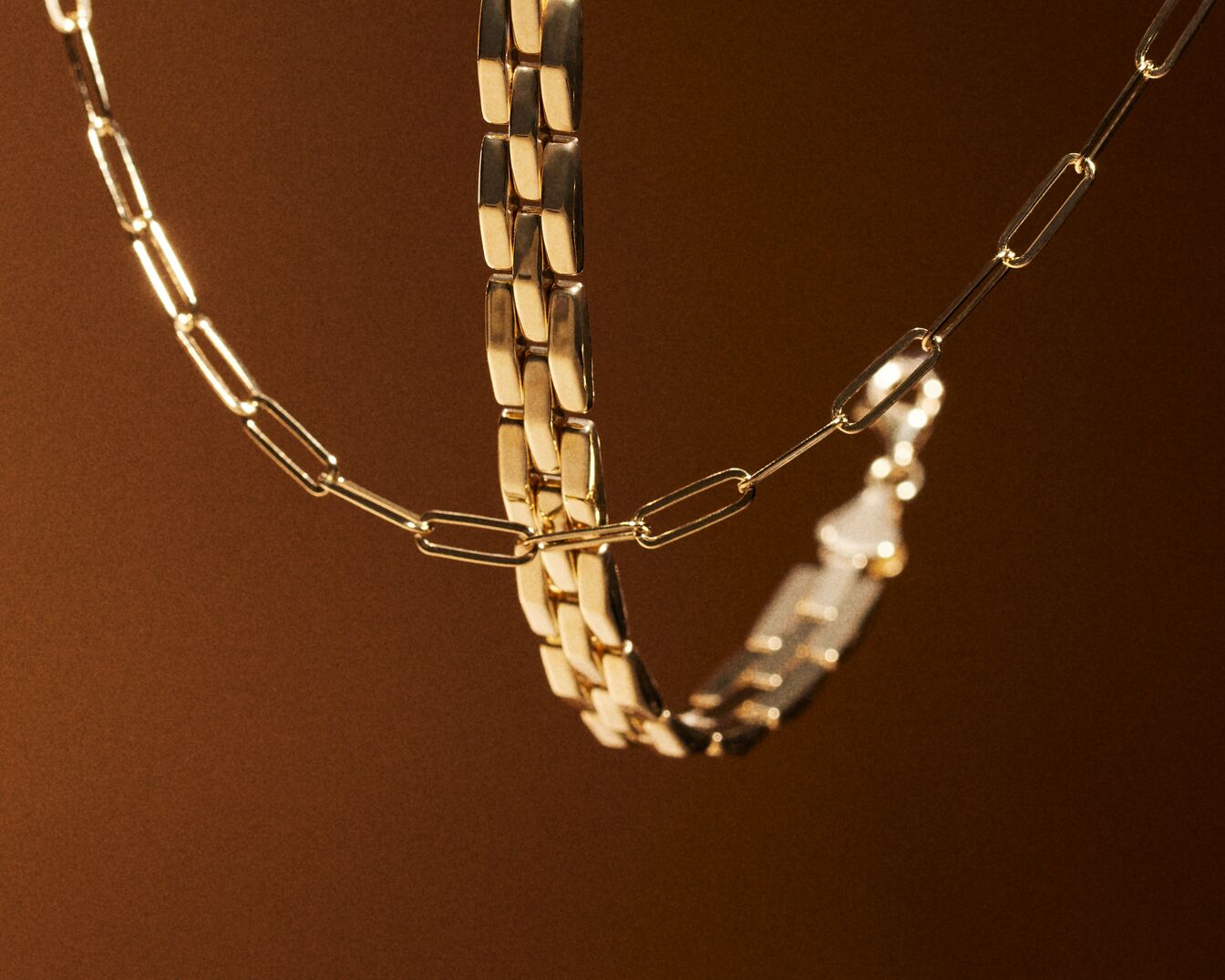 Twee gouden kettingen, een met rechthoekige schakels en een dikkere, platte ketting, hangen tegen een bruine achtergrond.