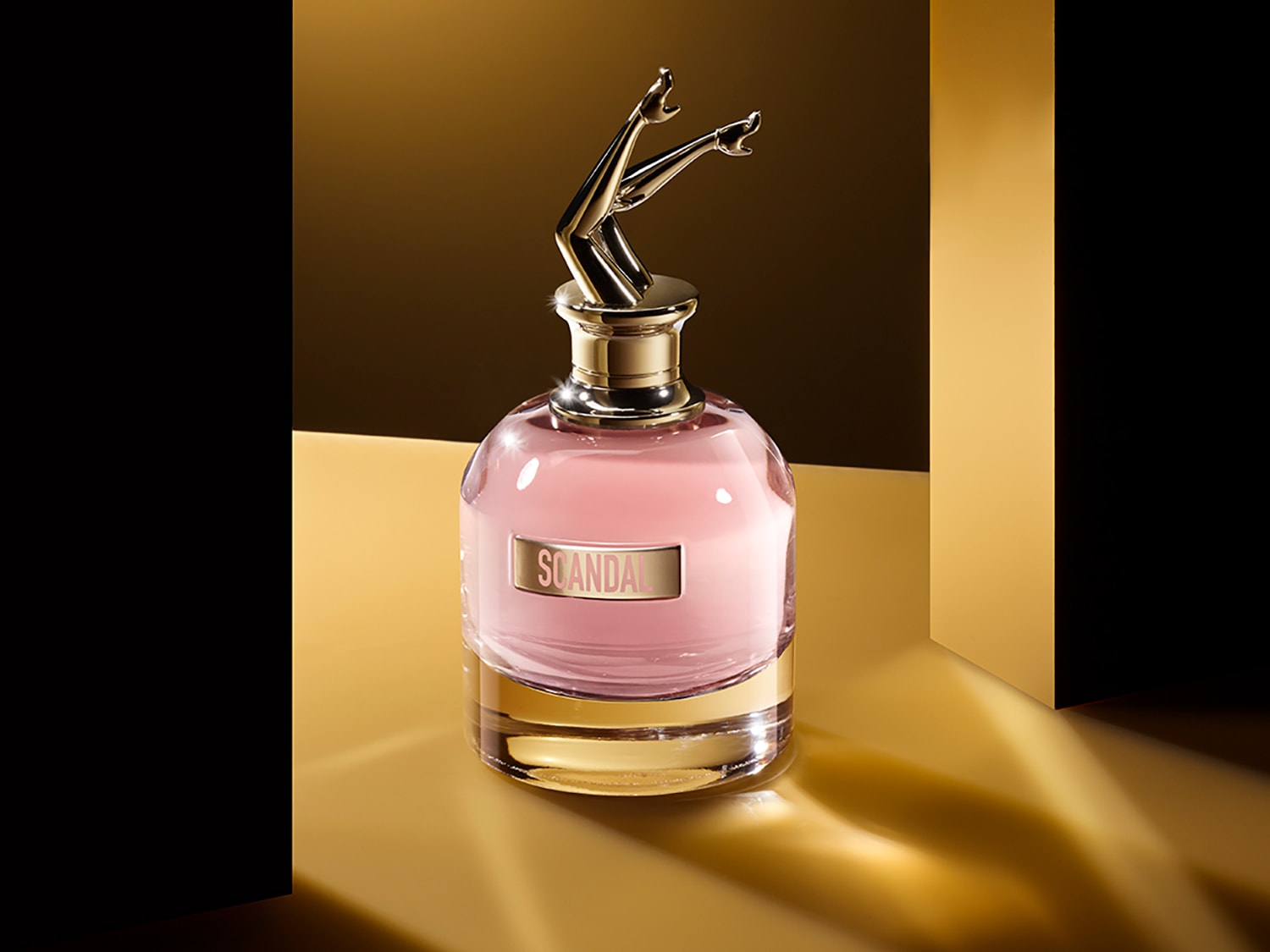 El frasco de perfume Scandal tiene un líquido rosado, una etiqueta dorada con el nombre y un tapón dorado con forma de piernas en el aire.