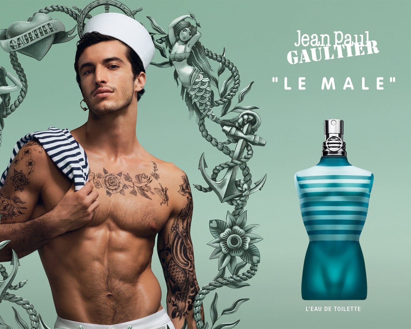 Un hombre con un sombrero de marinero blanco y un top de rayas azul marino y blanco sobre su hombro, mostrando tatuajes en el pecho y los brazos, posa junto a una botella de perfume Jean Paul Gaultier "Le Male" con forma de torso, con rayas horizontales blancas sobre un fondo azul verdoso.