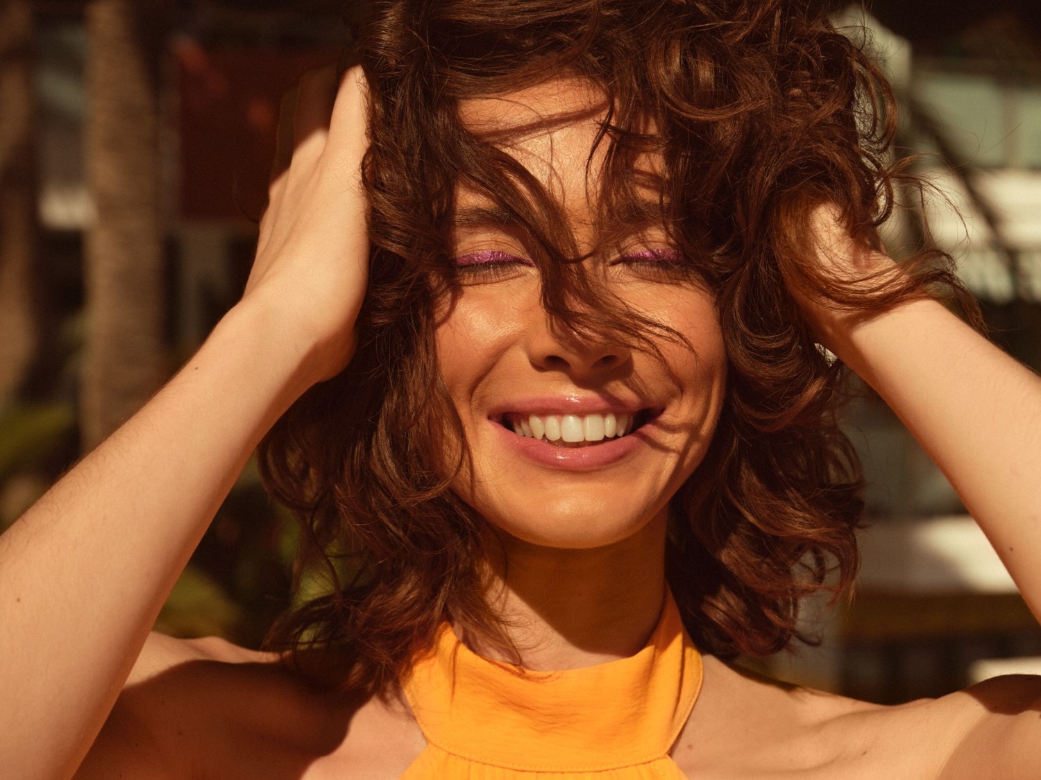 Een stralende vrouw met krullend bruin haar, paarse oogschaduw en een oranje top lacht terwijl ze haar handen in haar haar heeft.